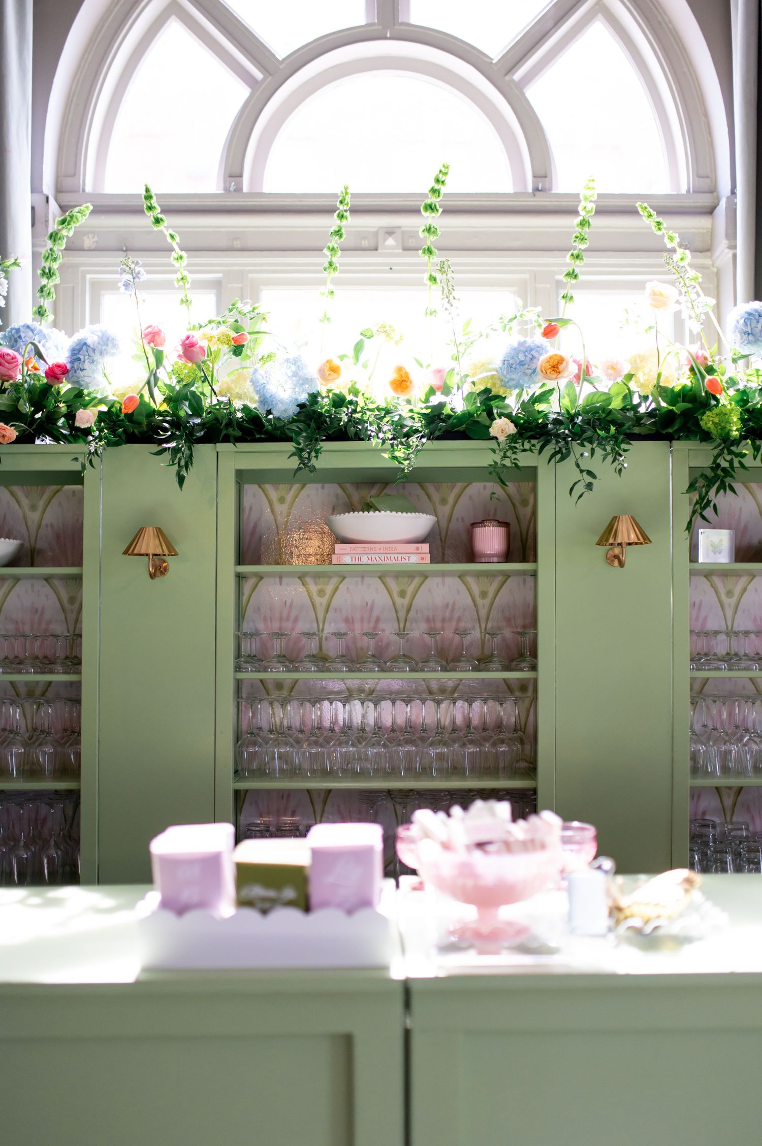 Green cabinet with floral garland, glassware, and pastel decor beneath an arched window in an elegant event space.