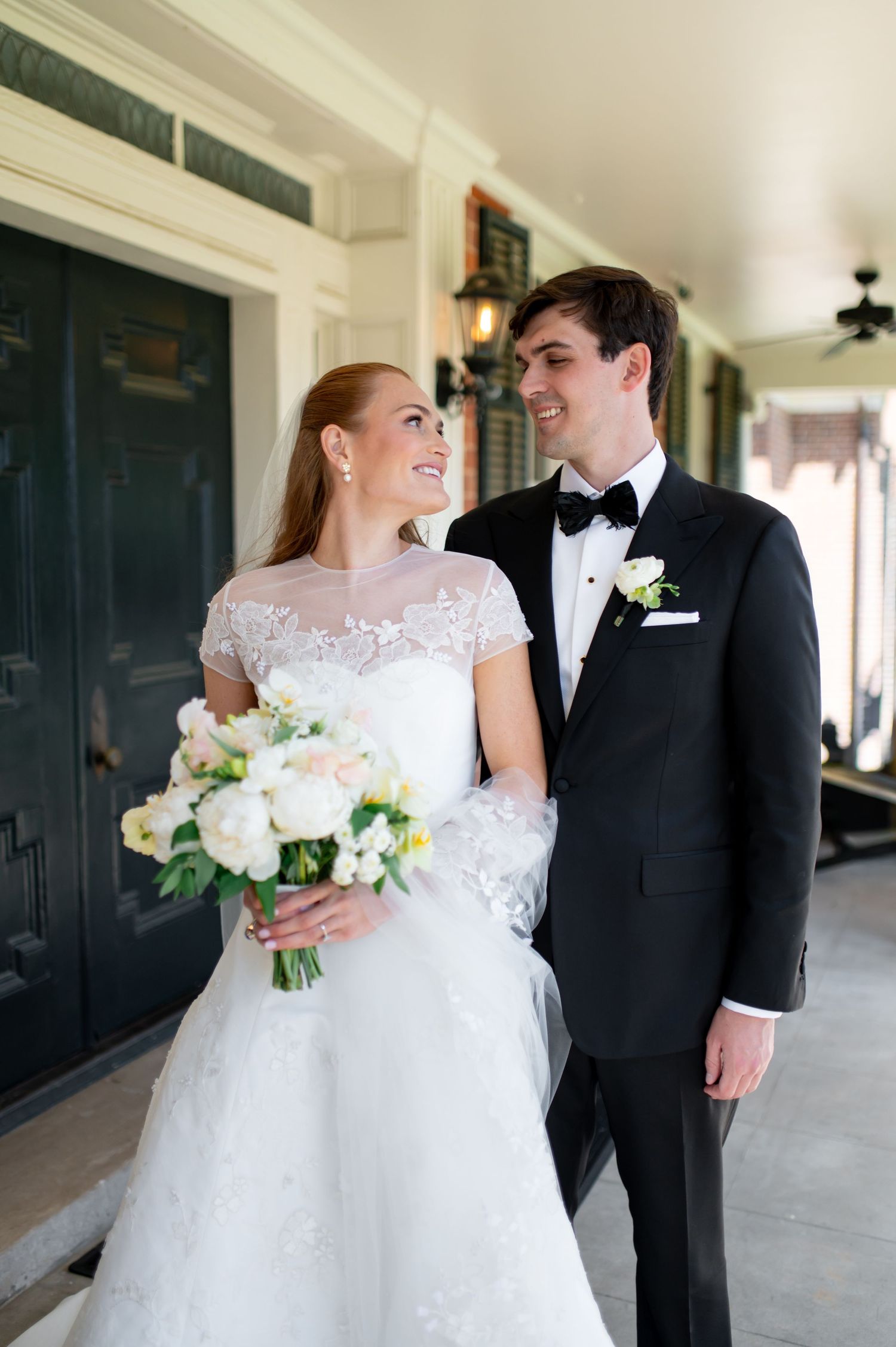 Smiling bride and groom sharing a loving glance on covered porch, bride holding white bouquet.