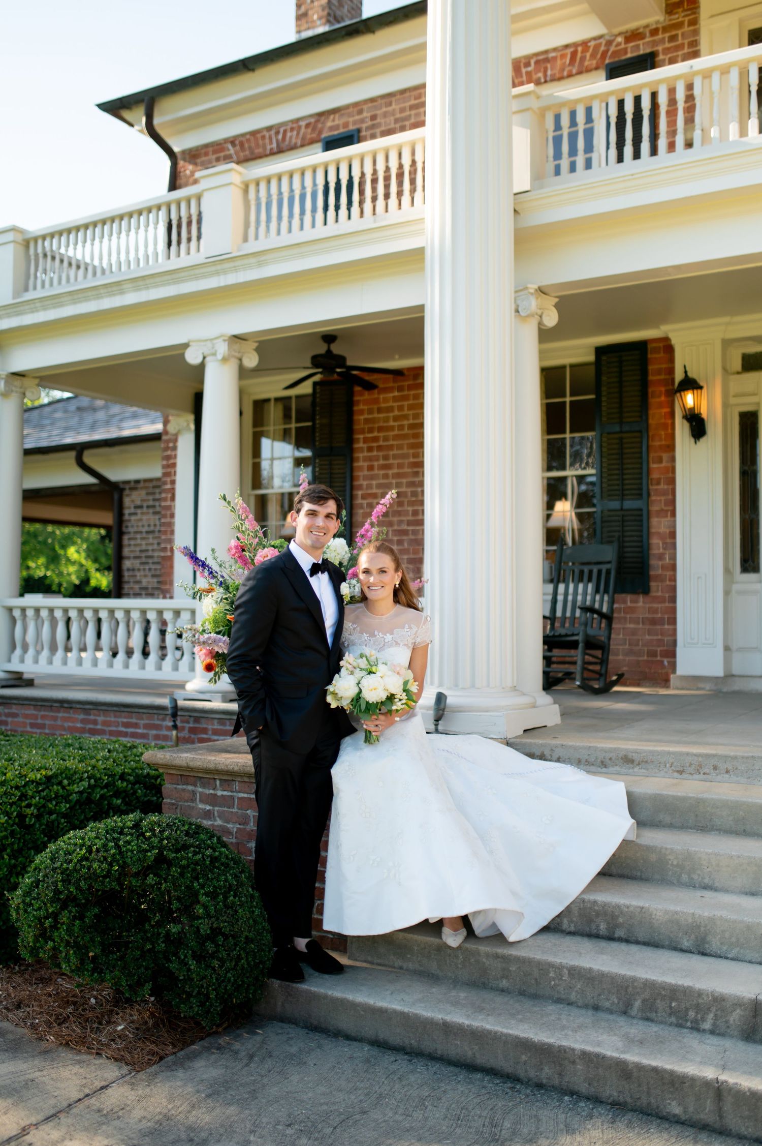 Bride and groom pose on steps of elegant Southern mansion with white columns at sunset.