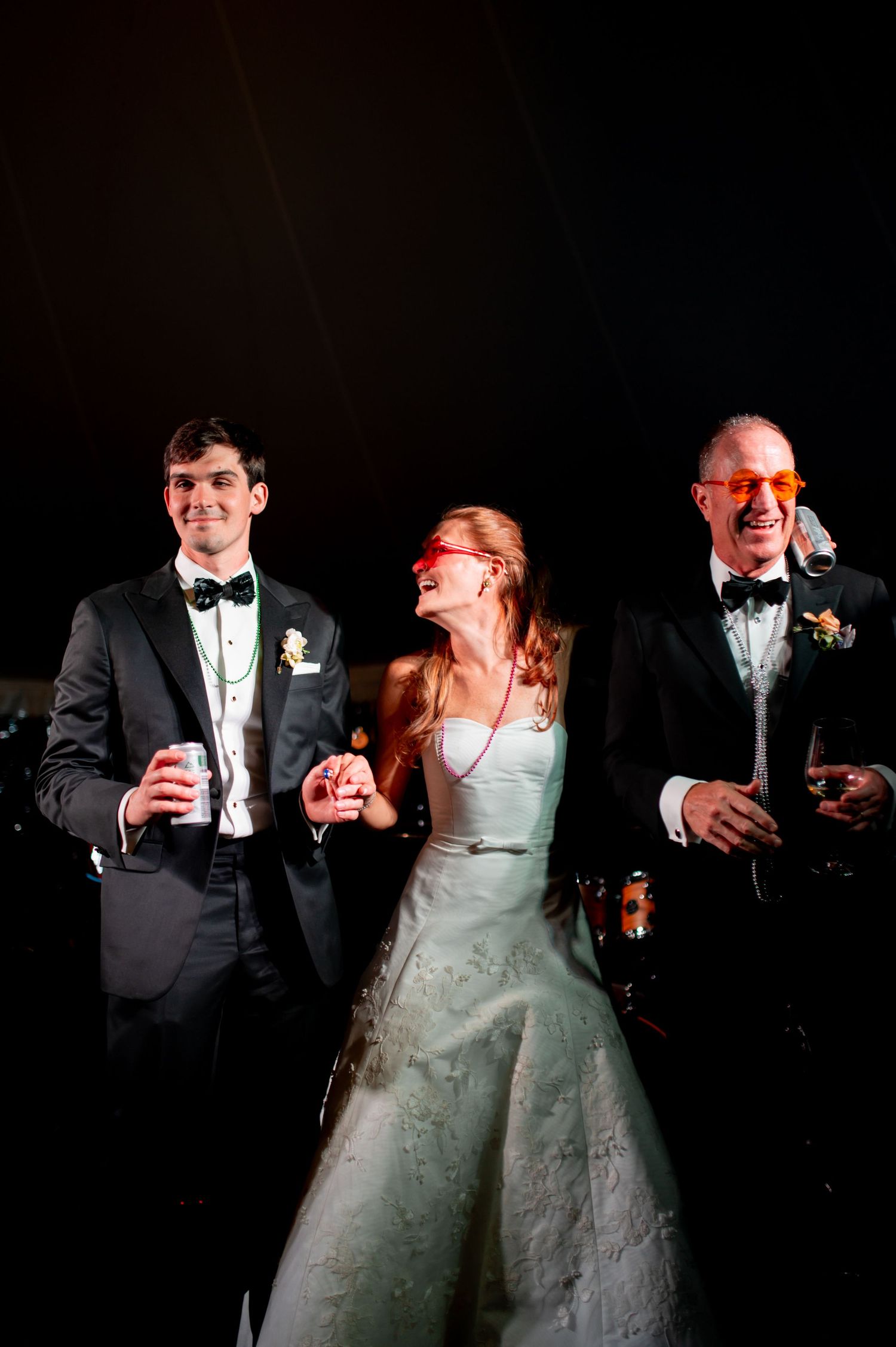Bride and groom laugh wearing novelty sunglasses with a guest during fun wedding reception celebration.