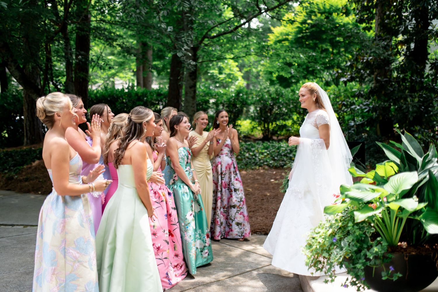 Bridesmaids in pastel floral gowns react with joy as bride in white dress and veil approaches in garden.