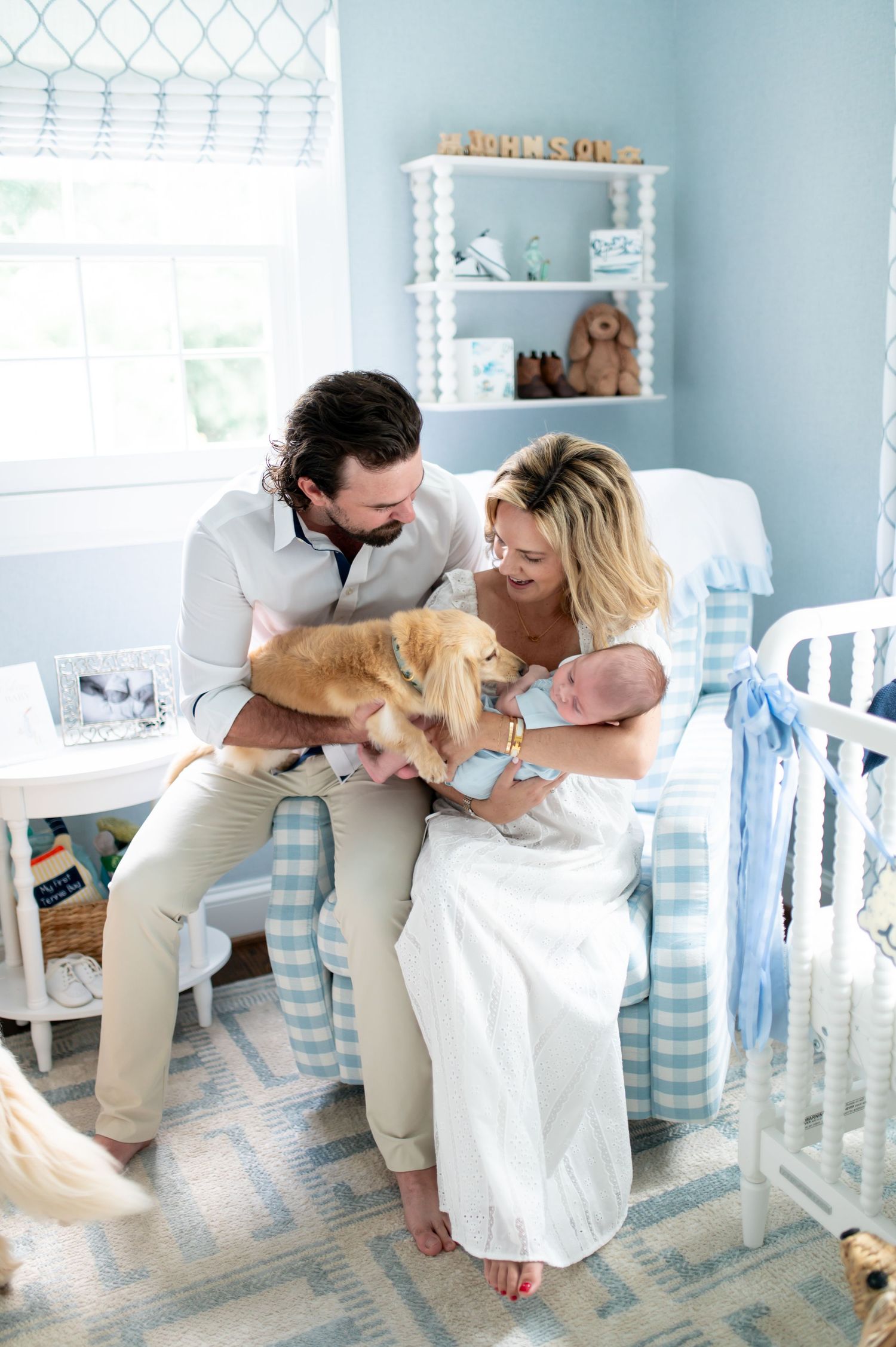 New parents sit in a blue nursery chair with their newborn baby and golden retriever dog.