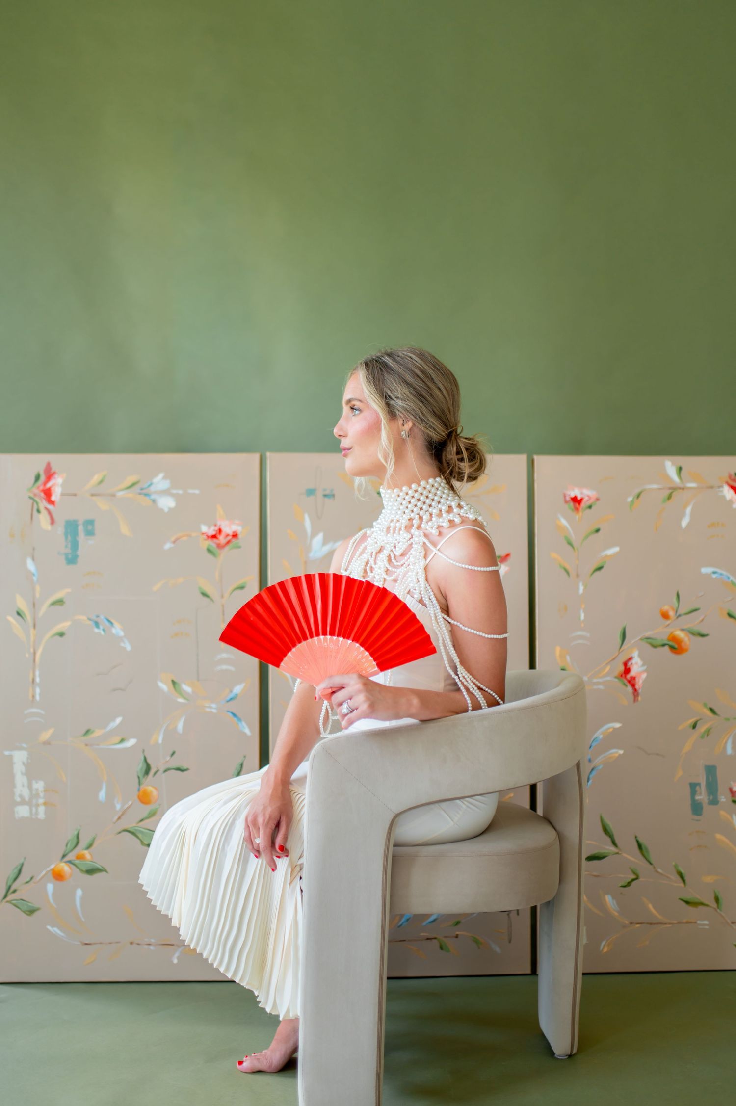 Elegant woman in white pearl dress holding red fan, seated in modern chair before green floral painted panels.
