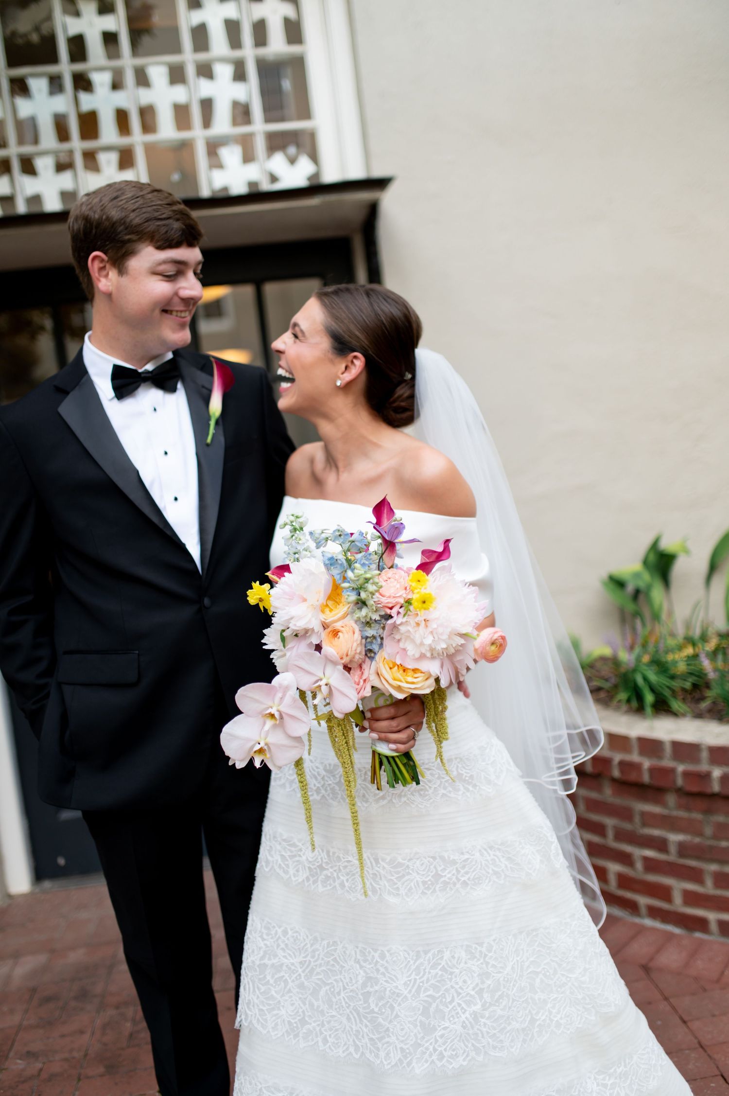 Joyful bride holding colorful spring bouquet laughs with groom in black tuxedo at outdoor wedding venue.