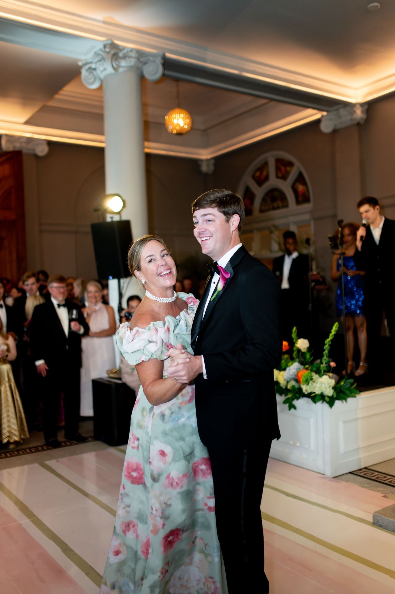 Mother and groom sharing a dance at wedding reception in grand ballroom with marble columns and floral centerpieces.