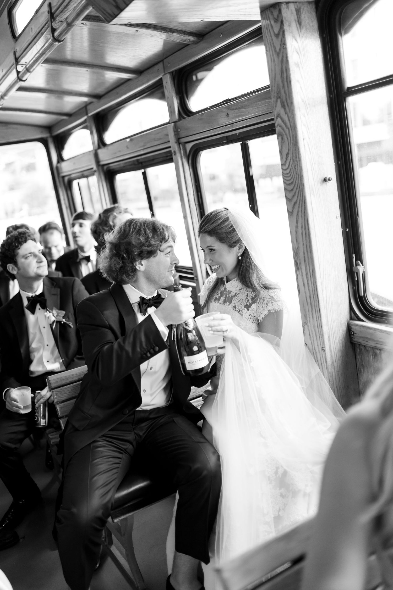 Black and white photo of newlyweds enjoying champagne on a trolley with wedding guests.