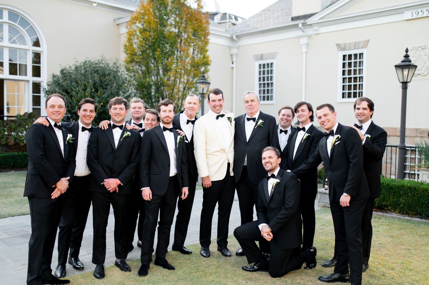 Wedding party in formal attire stands in front of a white colonial-style building.