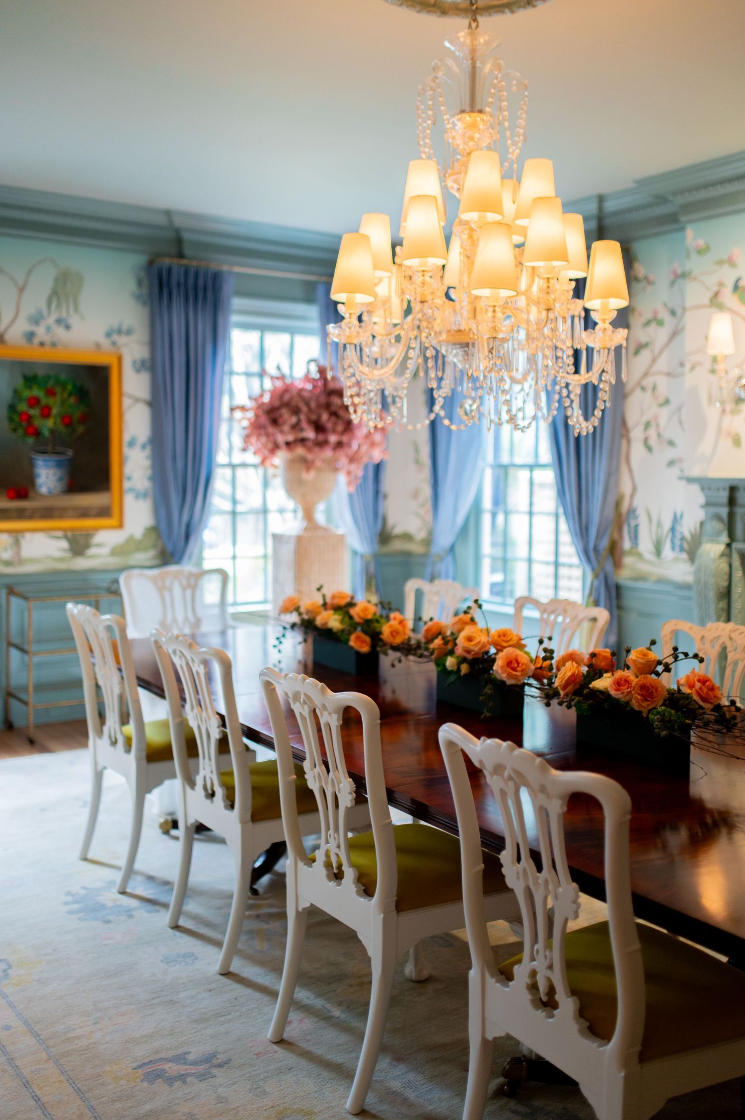 Elegant blue dining room with crystal chandelier, white Chippendale chairs, dark wood table, and orange floral centerpiece.