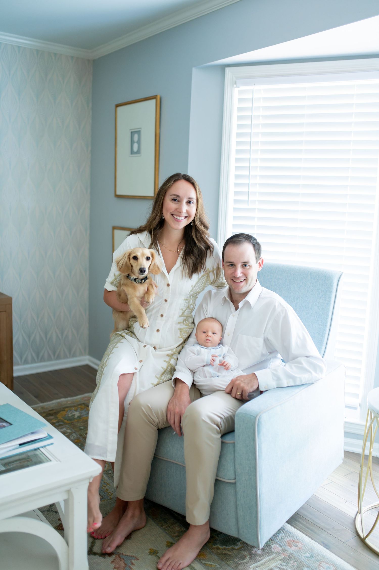 Young family with newborn baby and golden dachshund poses in a soft blue nursery room.