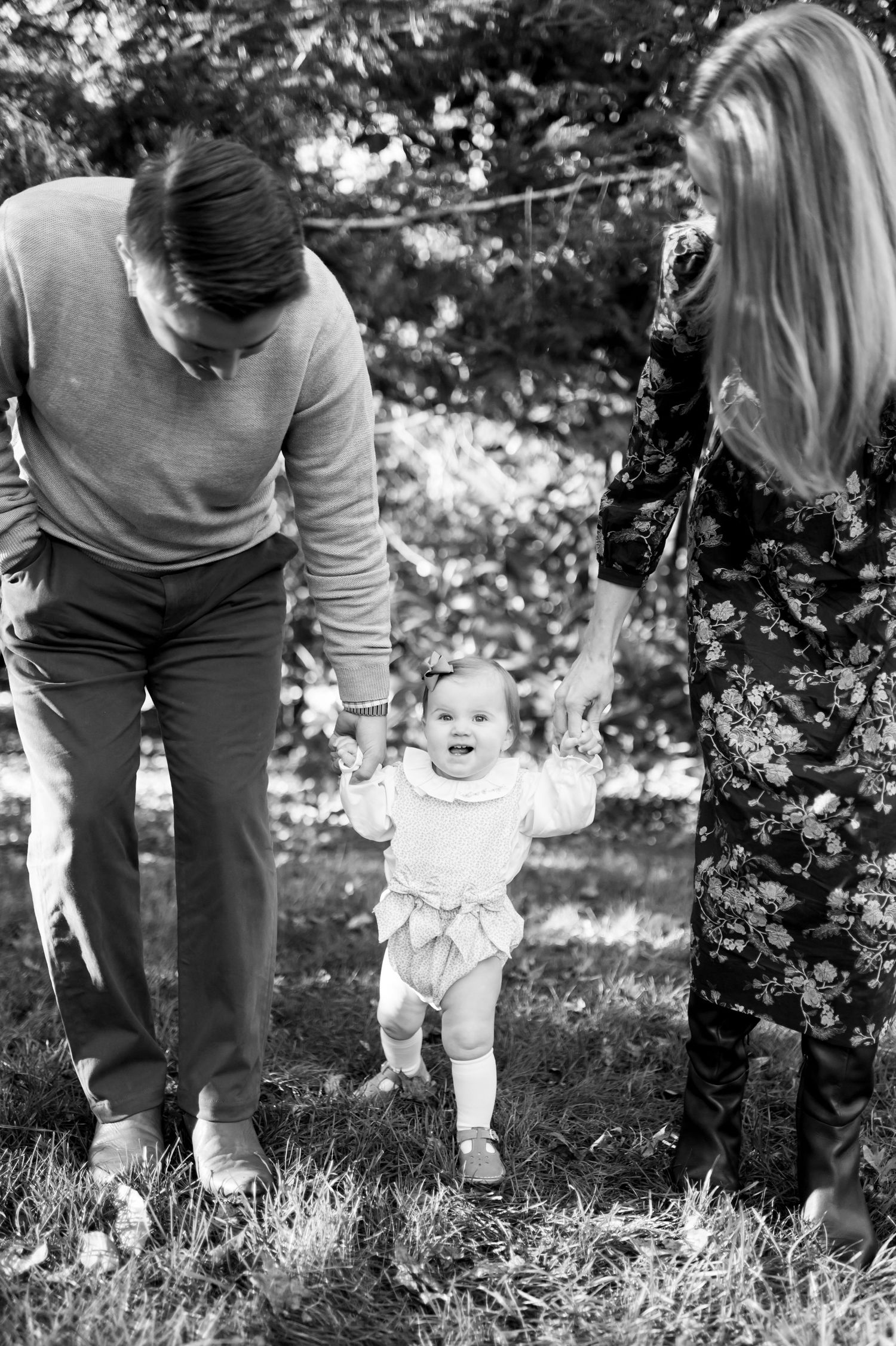 Black and white photo of parents helping their smiling baby take first steps outdoors in a sunny garden.