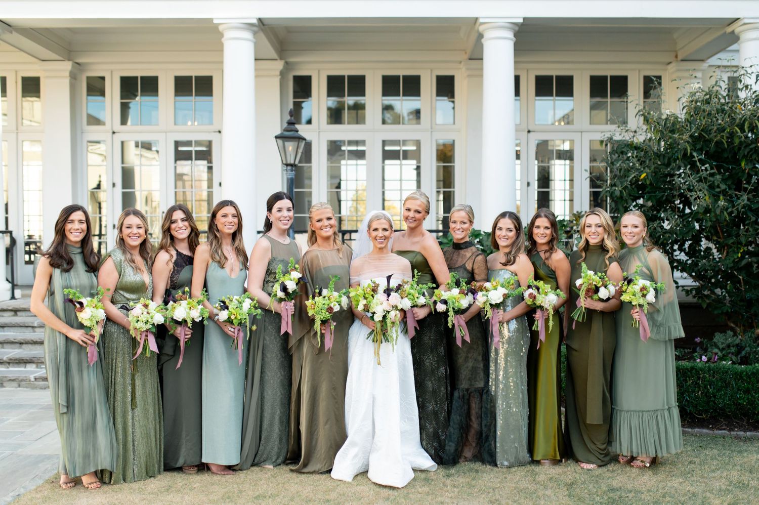 Wedding party in sage green dresses and white gown pose in front of columned building.