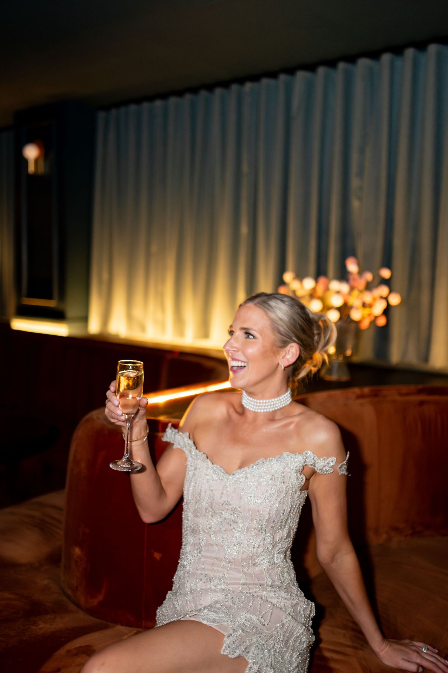 Bride in lace dress laughing while seated on brown leather couch.