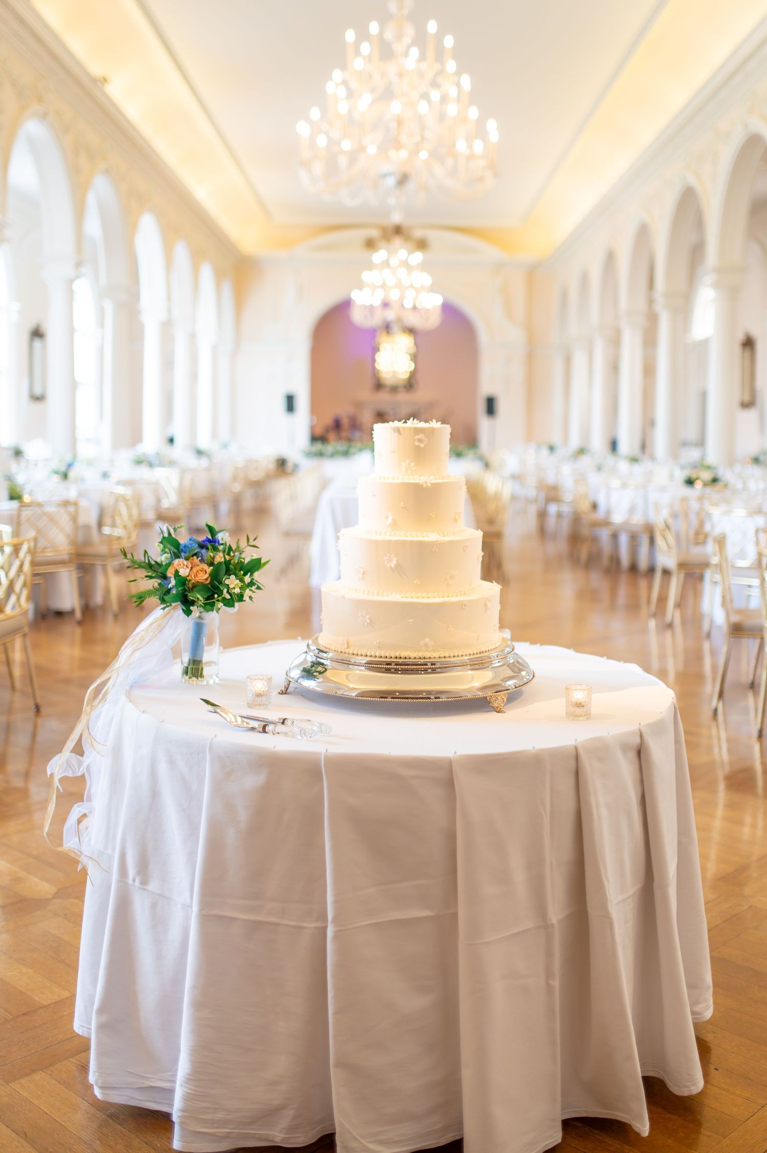 White wedding cake displayed on round table in luxurious ballroom with chandeliers.