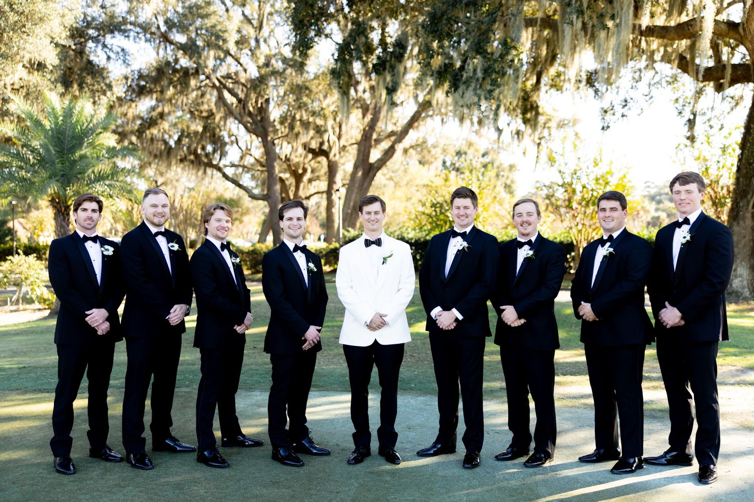 Groomsmen in black tuxedos and bowties stand in a line outdoors for a formal wedding photo.