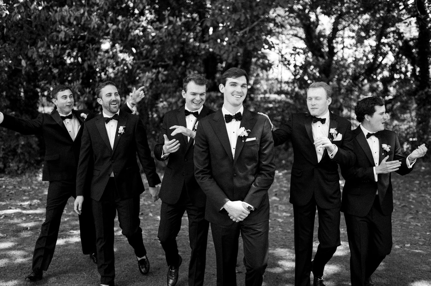 Groom and groomsmen in black tuxedos laughing and walking together outdoors in black and white photo.