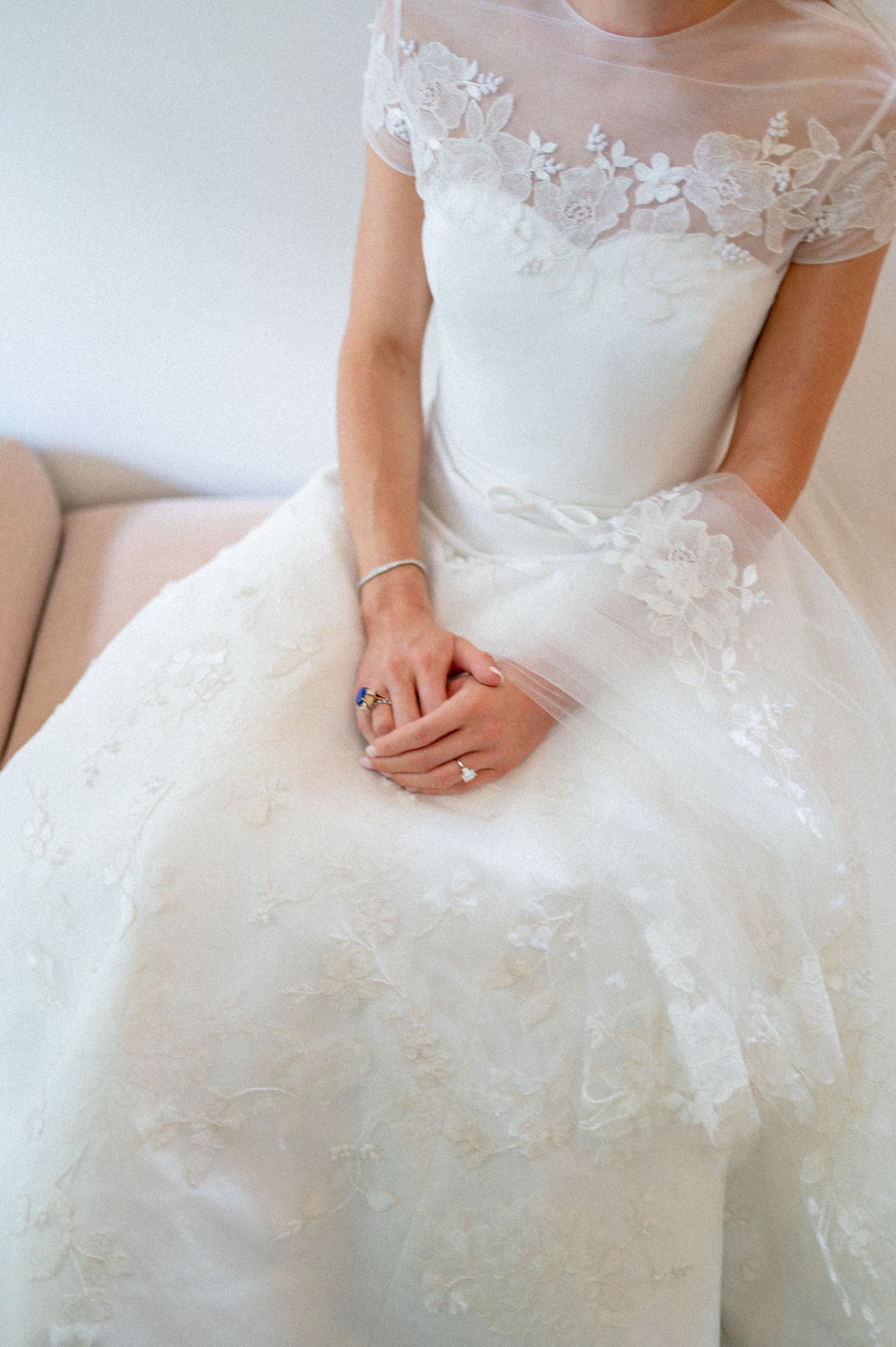 Close-up of bride in floral lace wedding gown with cap sleeves, hands folded in lap showing engagement ring.