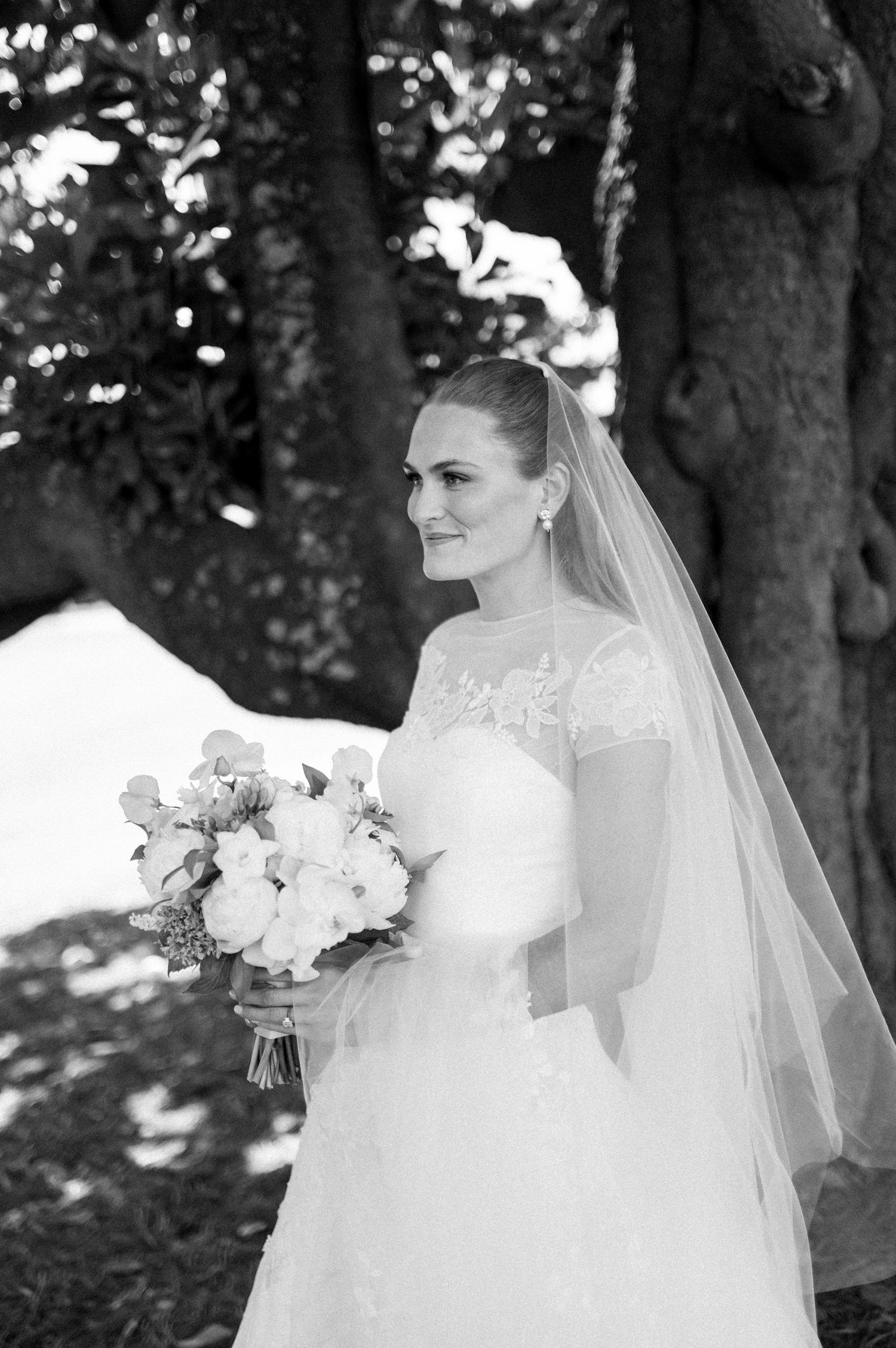 Black and white portrait of bride in lace veil holding bouquet under sunlit trees.