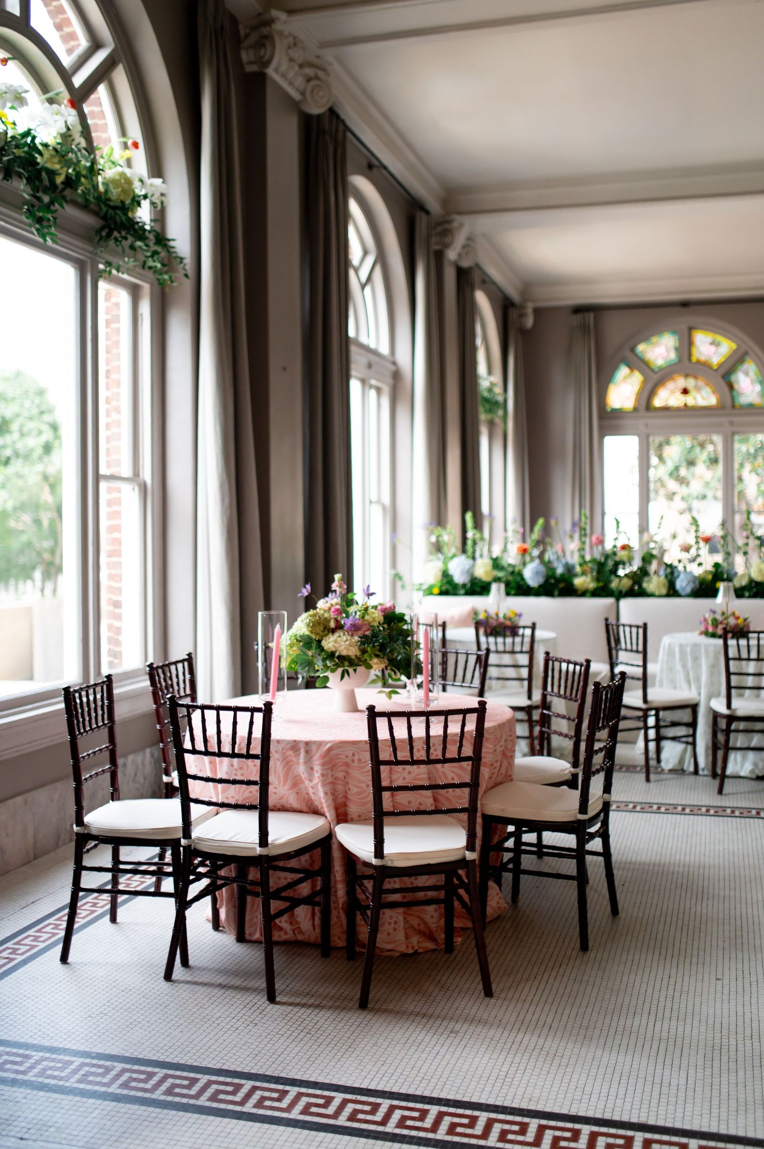Wedding reception hall with arched windows, pink tablecloths, chiavari chairs, and floral centerpieces.