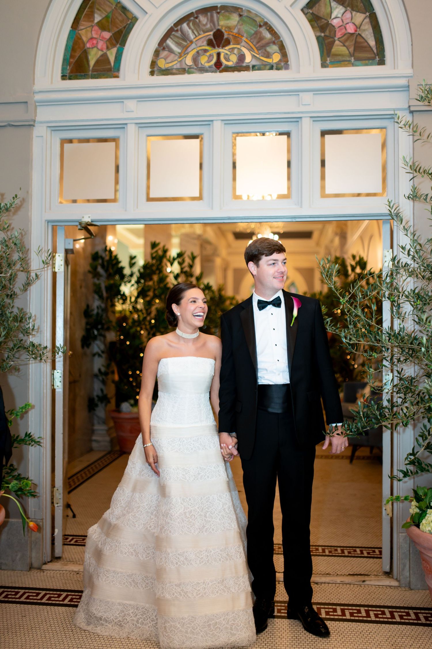 Smiling bride and groom holding hands at ornate venue entrance with stained glass transom and lush greenery decor.