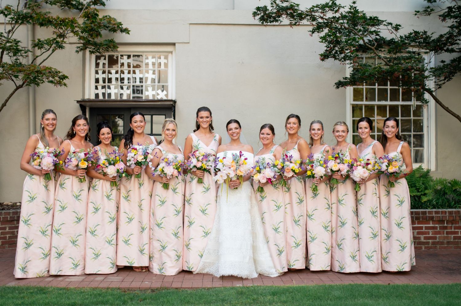 Bride and bridesmaids in floral print gowns holding colorful bouquets pose together outdoors at elegant wedding venue.