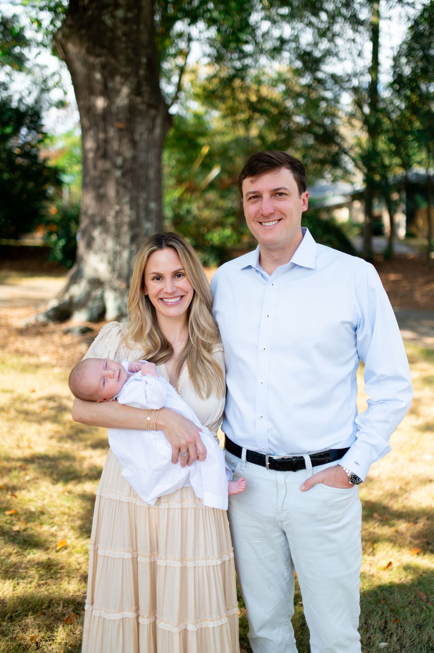 A couple in coordinated light-colored outfits poses outdoors with their newborn baby under a large tree.