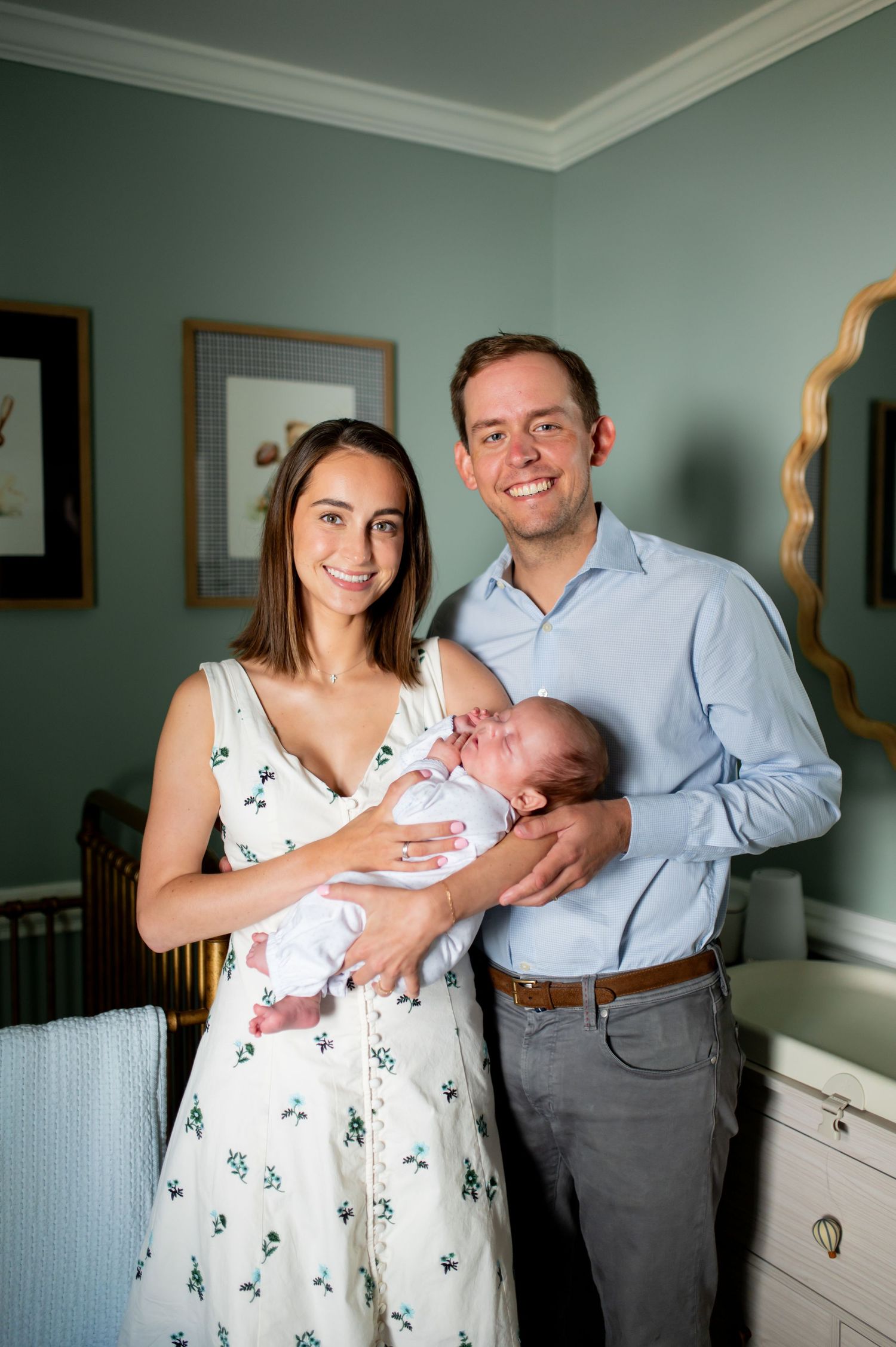 A couple holds their newborn baby in a mint green nursery room with white furniture.