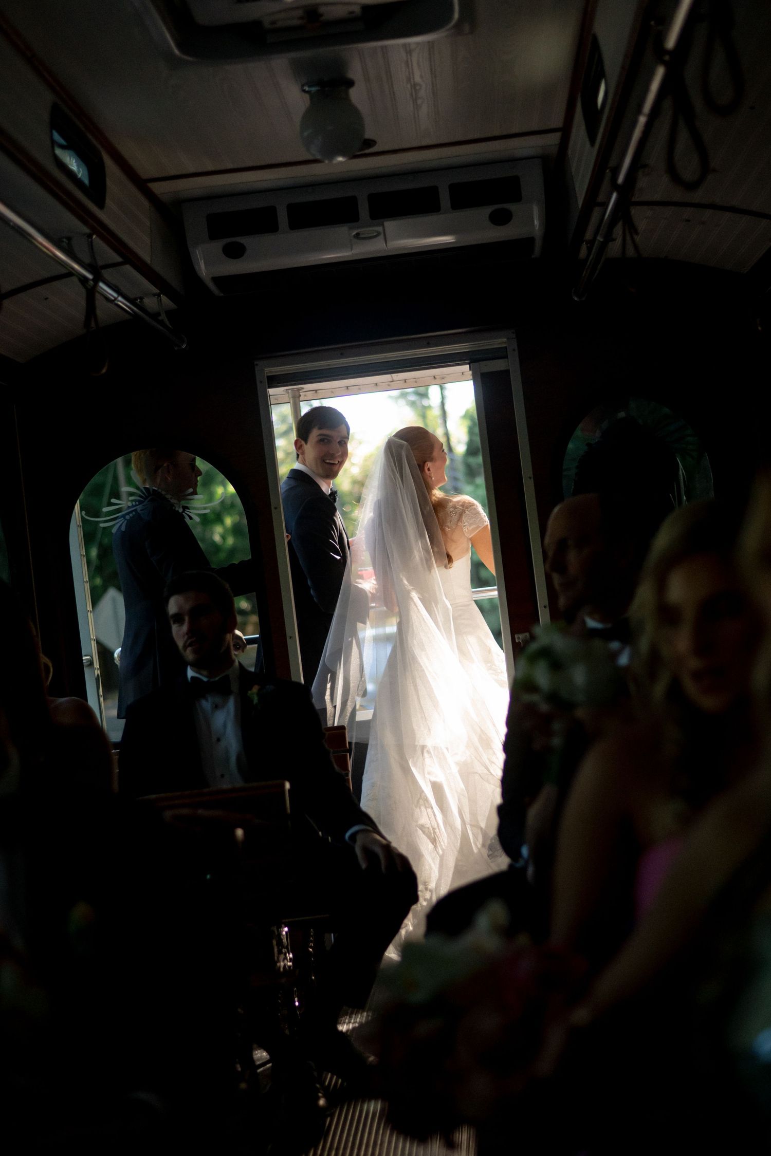Bride in white gown and groom standing at trolley door with wedding guests seated inside.