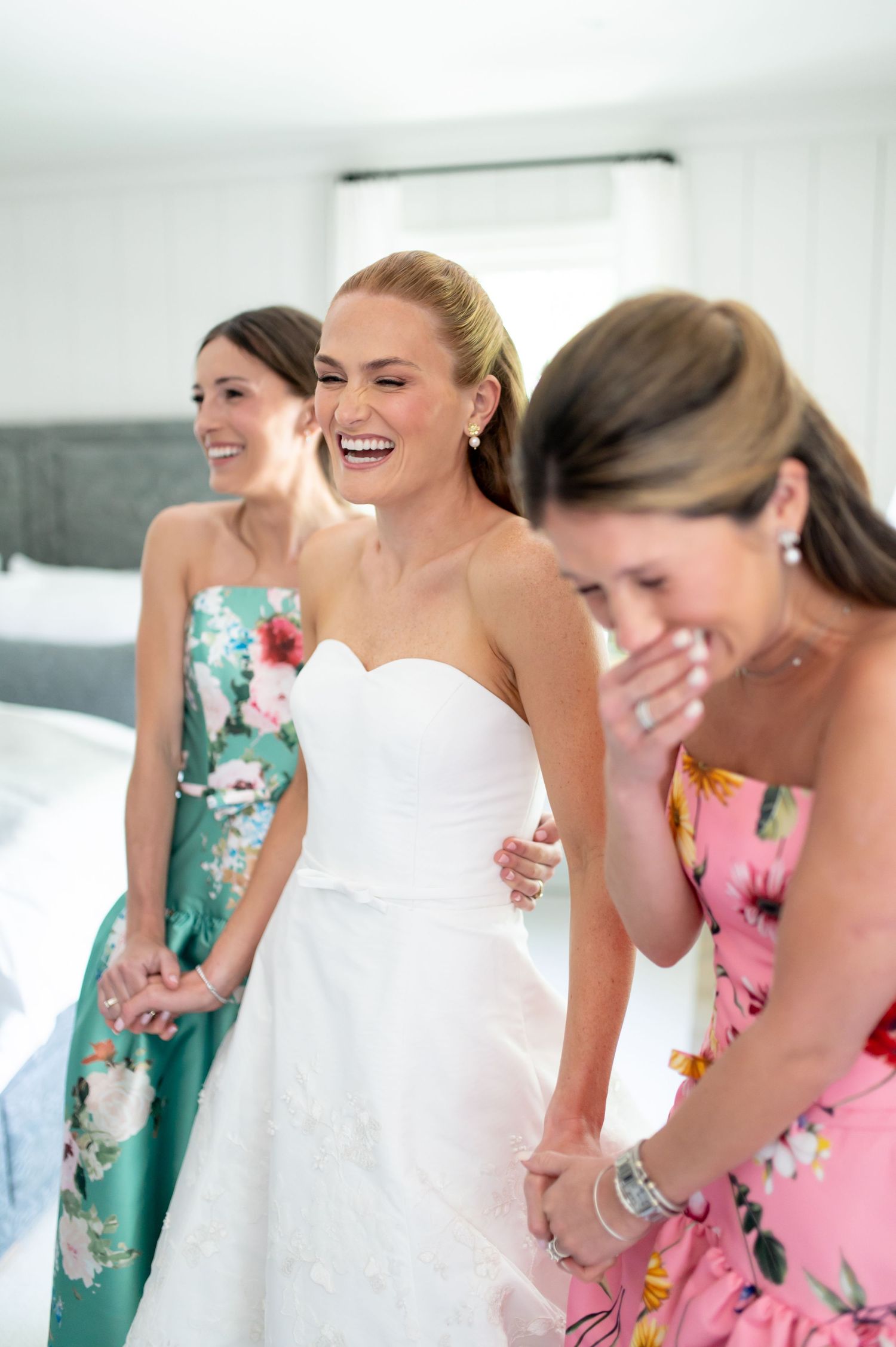 Bride in white strapless gown laughing joyfully with bridesmaids in colorful floral dresses during wedding preparations.