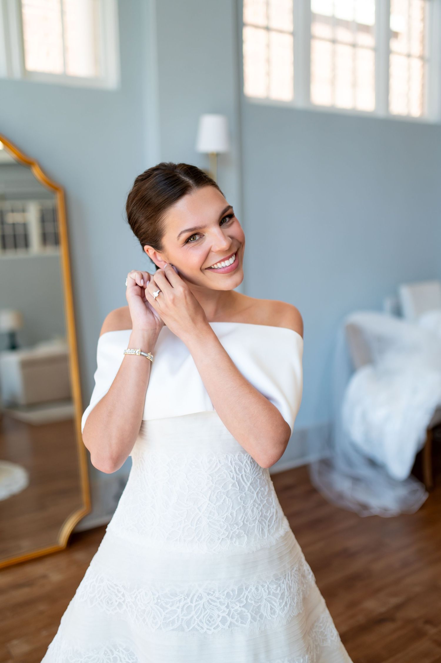 Smiling bride in off-shoulder white gown putting on earrings in a bright room with gold mirror.