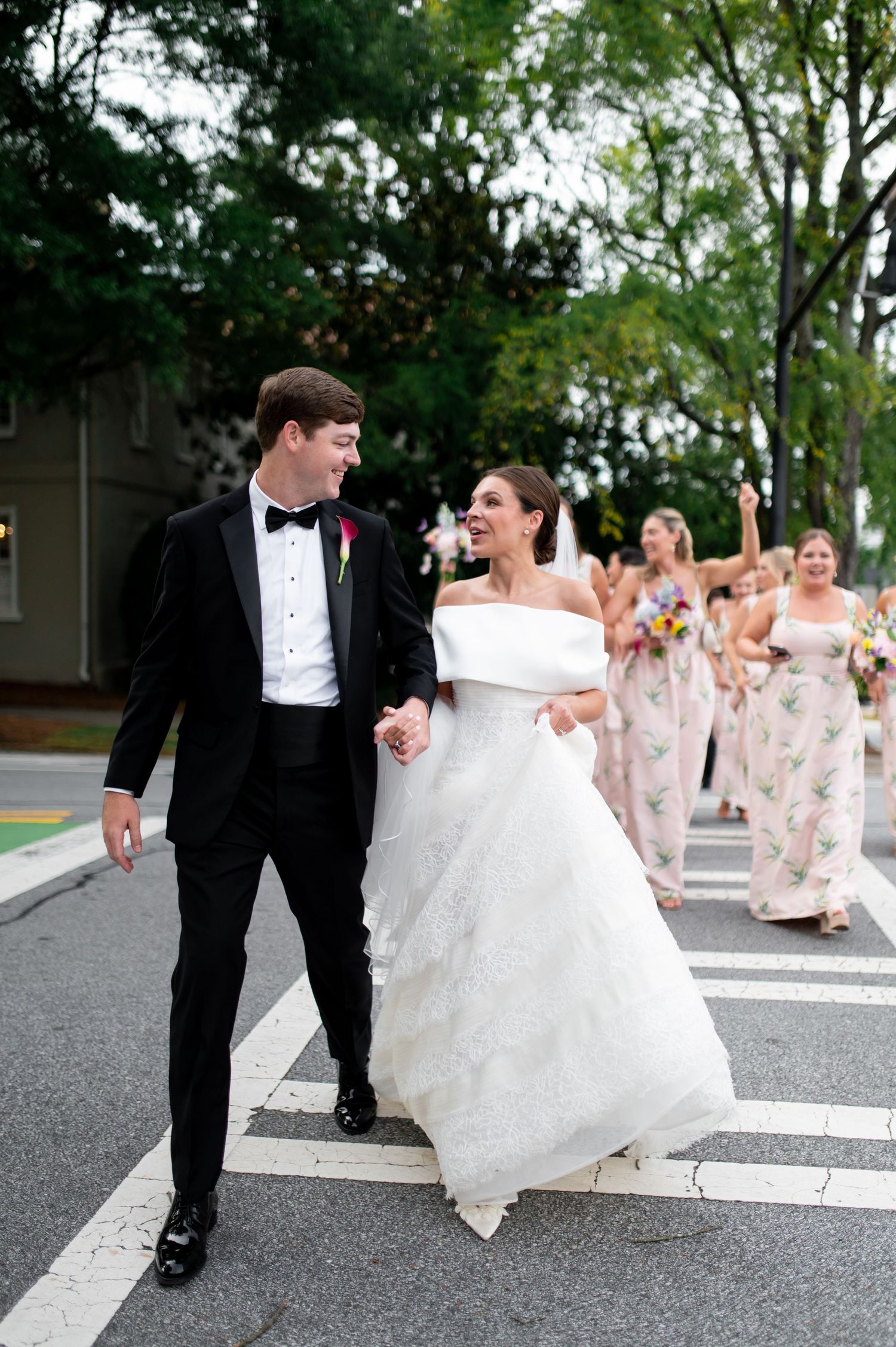 Newlyweds cross street hand-in-hand, bridesmaids in pink floral dresses celebrating behind them.