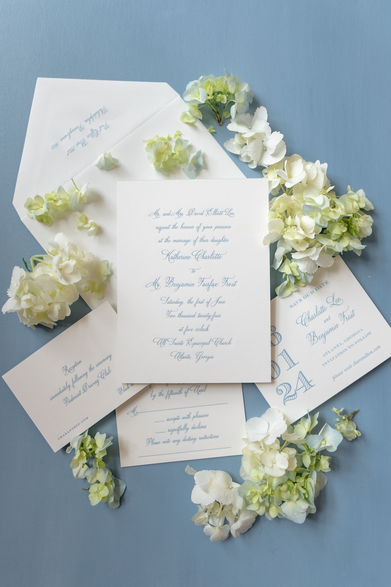 Wedding stationery suite with blue script and white hydrangea blooms on pale blue surface.