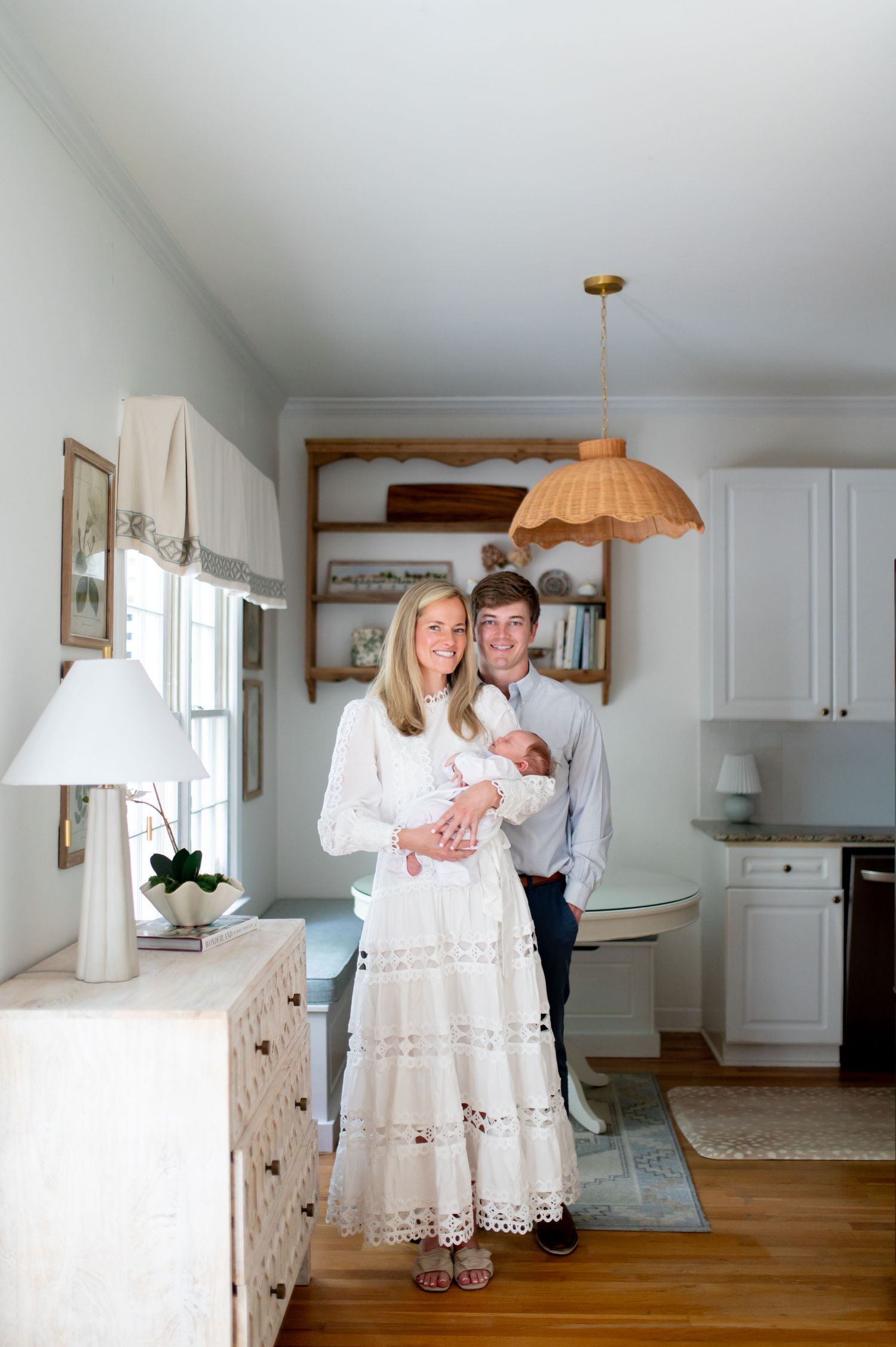 A smiling couple in a bright home holds their newborn baby, the woman in a white lace dress and man in navy attire.