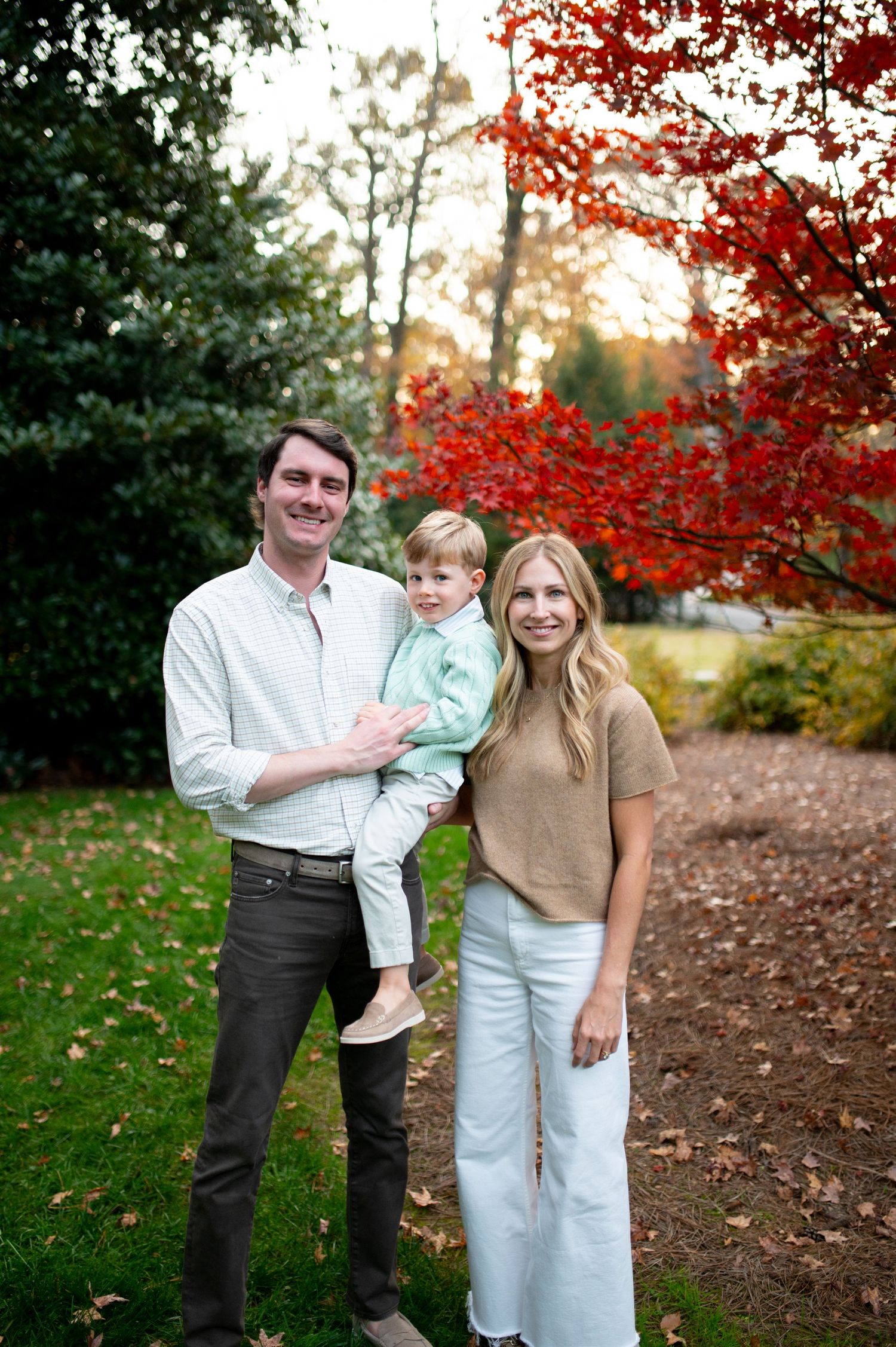 Family of three posing outdoors in autumn garden with vibrant red maple tree and green lawn backdrop.