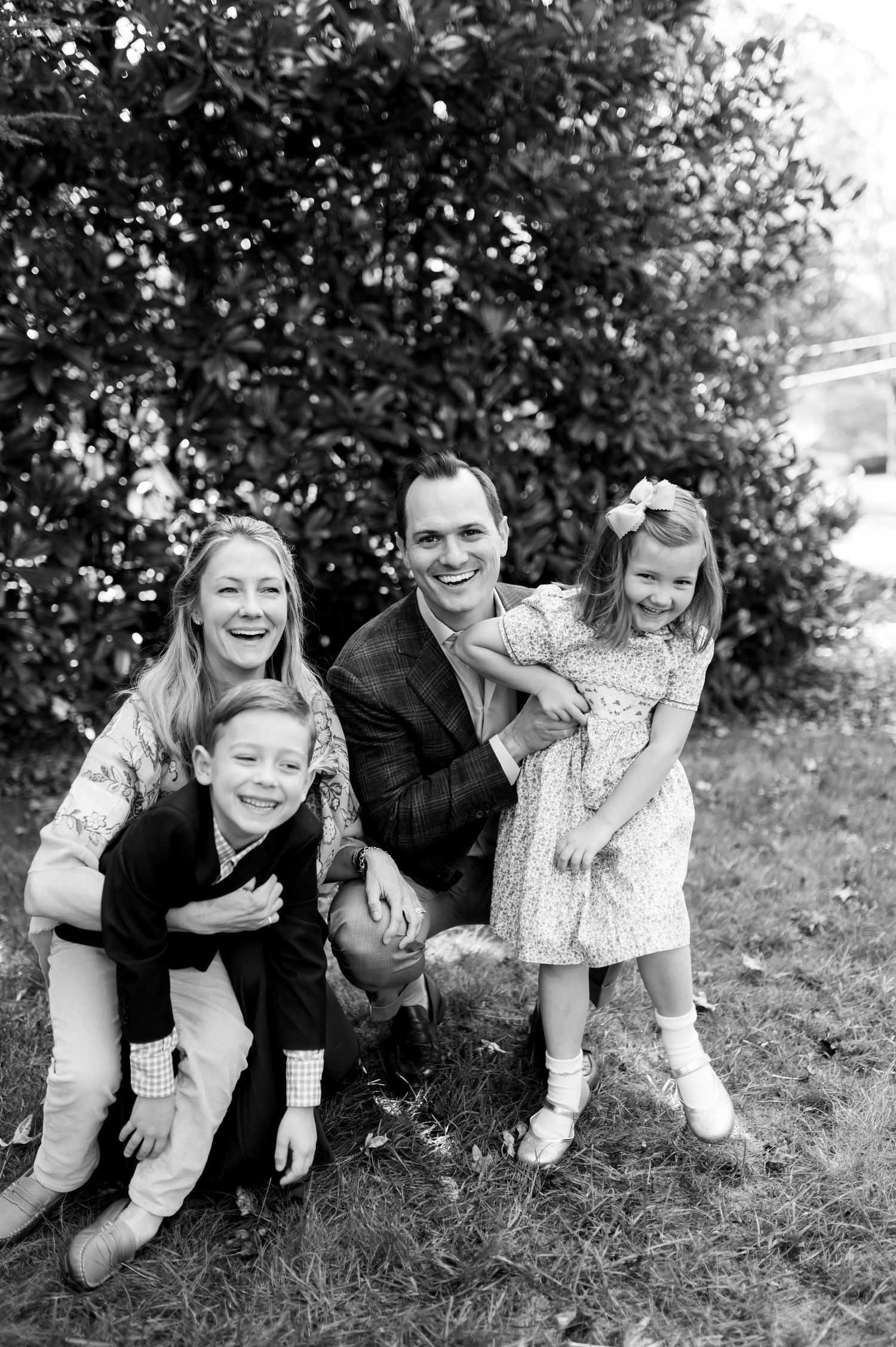 Black and white family portrait of parents and two young children laughing outdoors near a lush green bush.