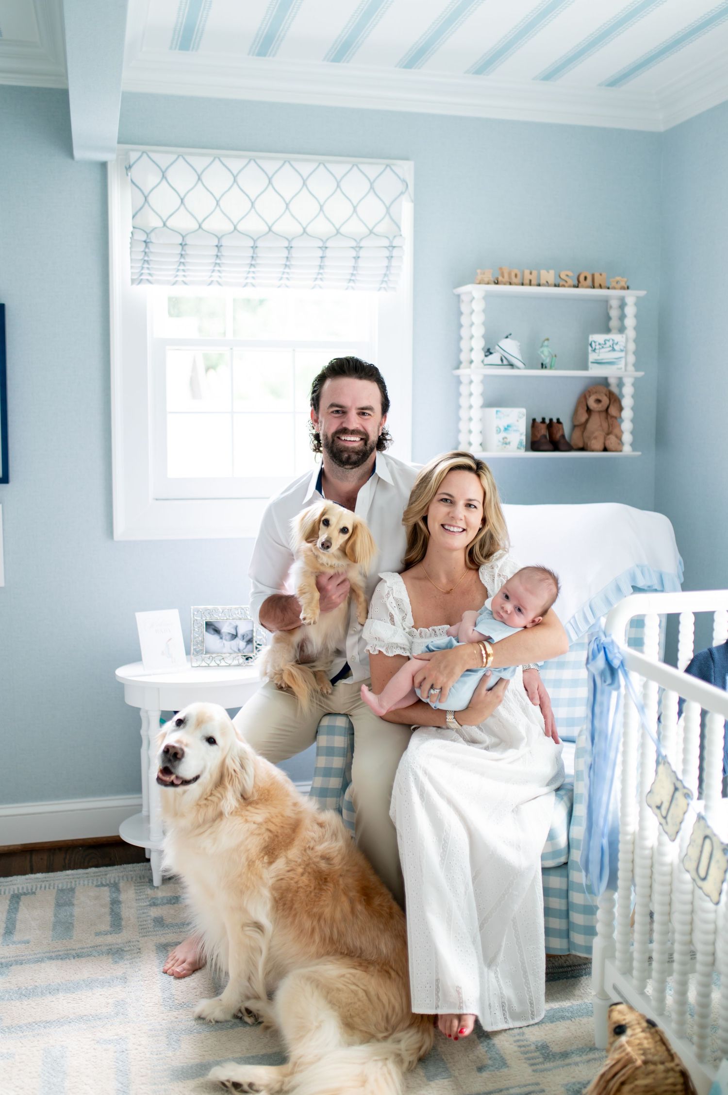 Happy family with newborn baby, two dogs pose in a soft blue nursery room with white crib and shelves.