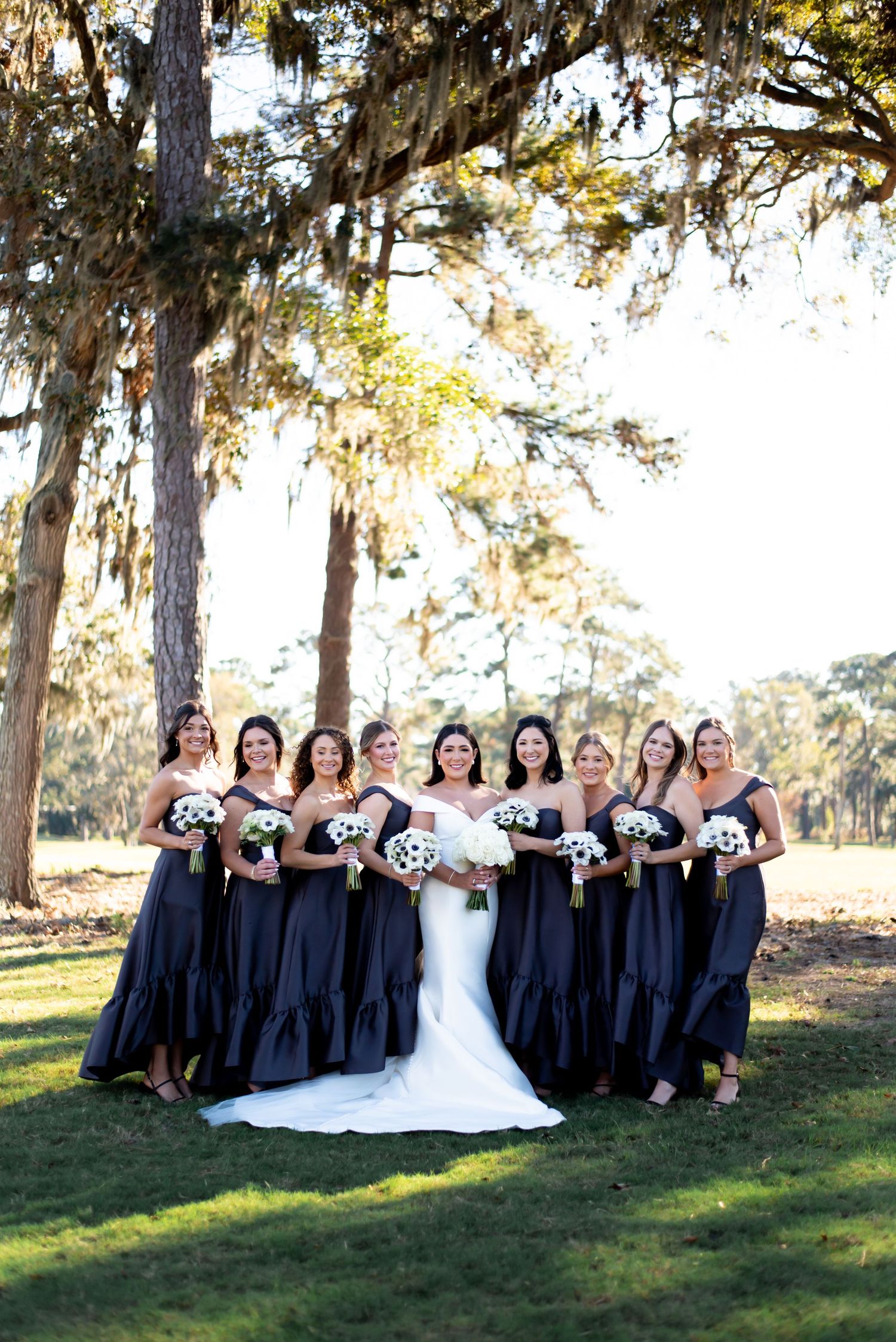 Wedding party in navy dresses and white gown pose together on grass under tall trees.