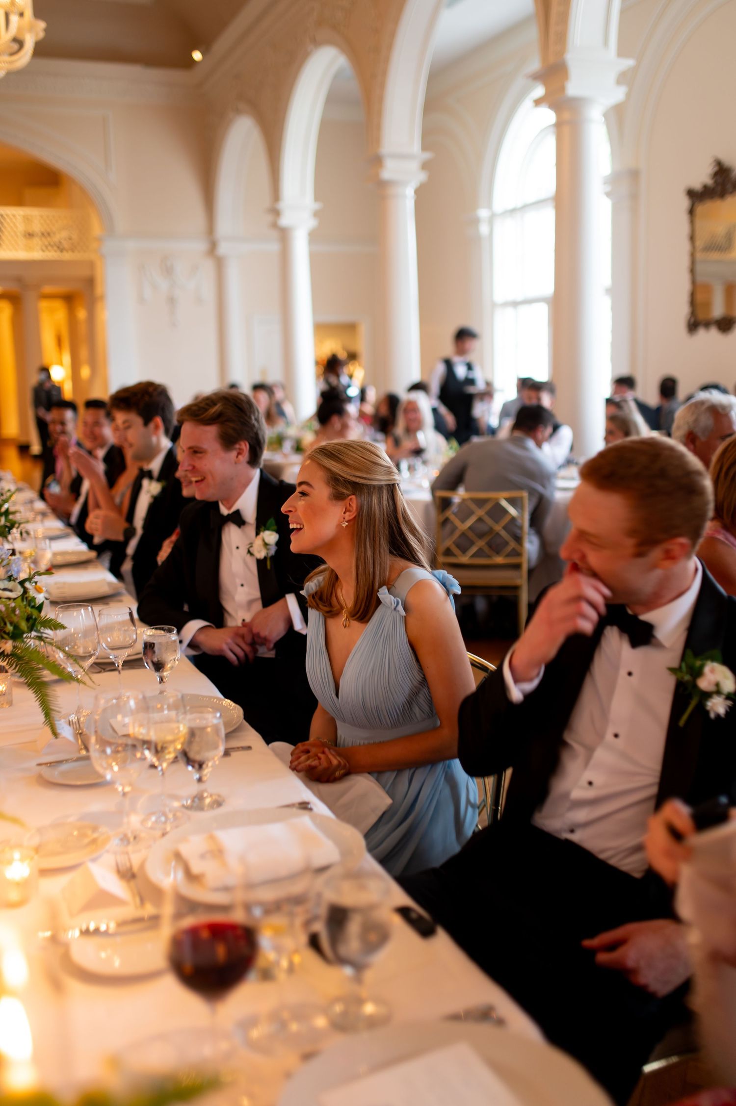 Wedding reception guests seated at elegant tables in a grand ballroom with chandeliers and arched windows.
