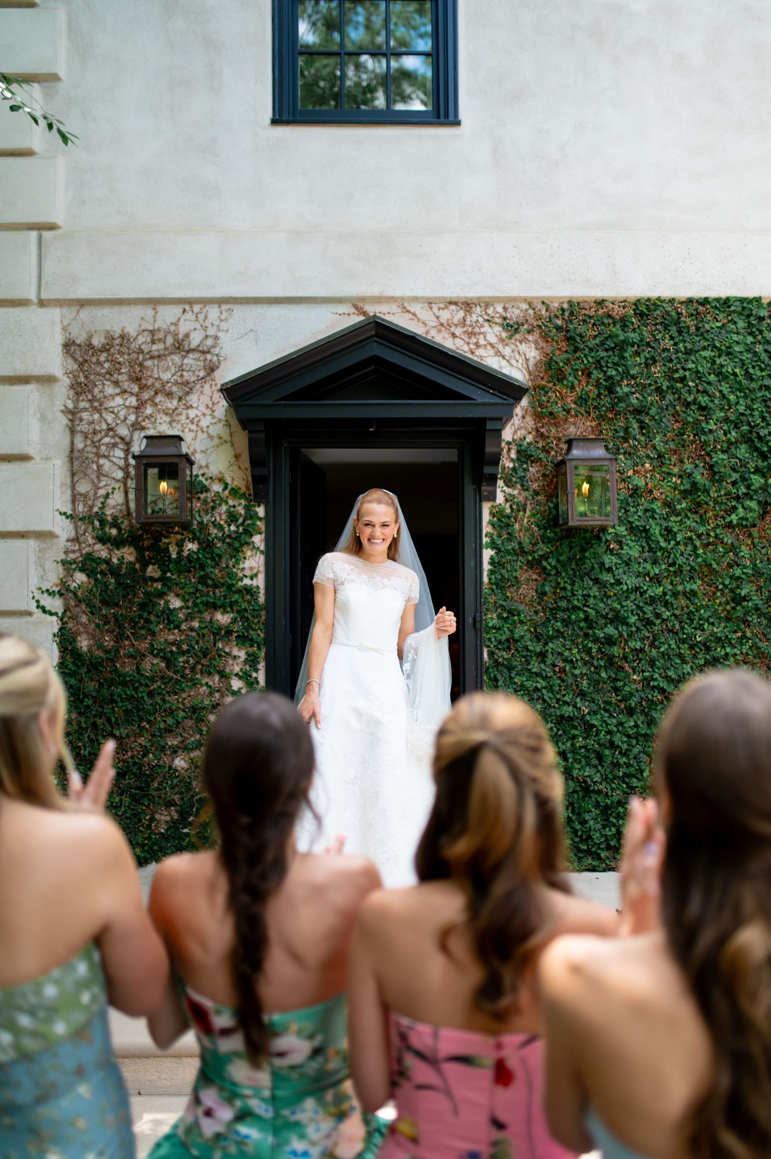 Bride in white gown and veil smiles at cheering bridesmaids outside ivy-covered venue entrance.