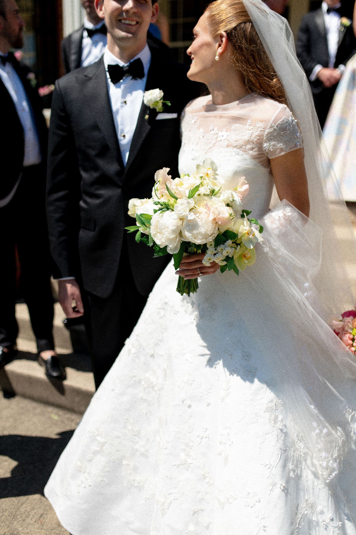 Bride holding white peony bouquet in lace cap sleeve gown beside groom in black tuxedo.
