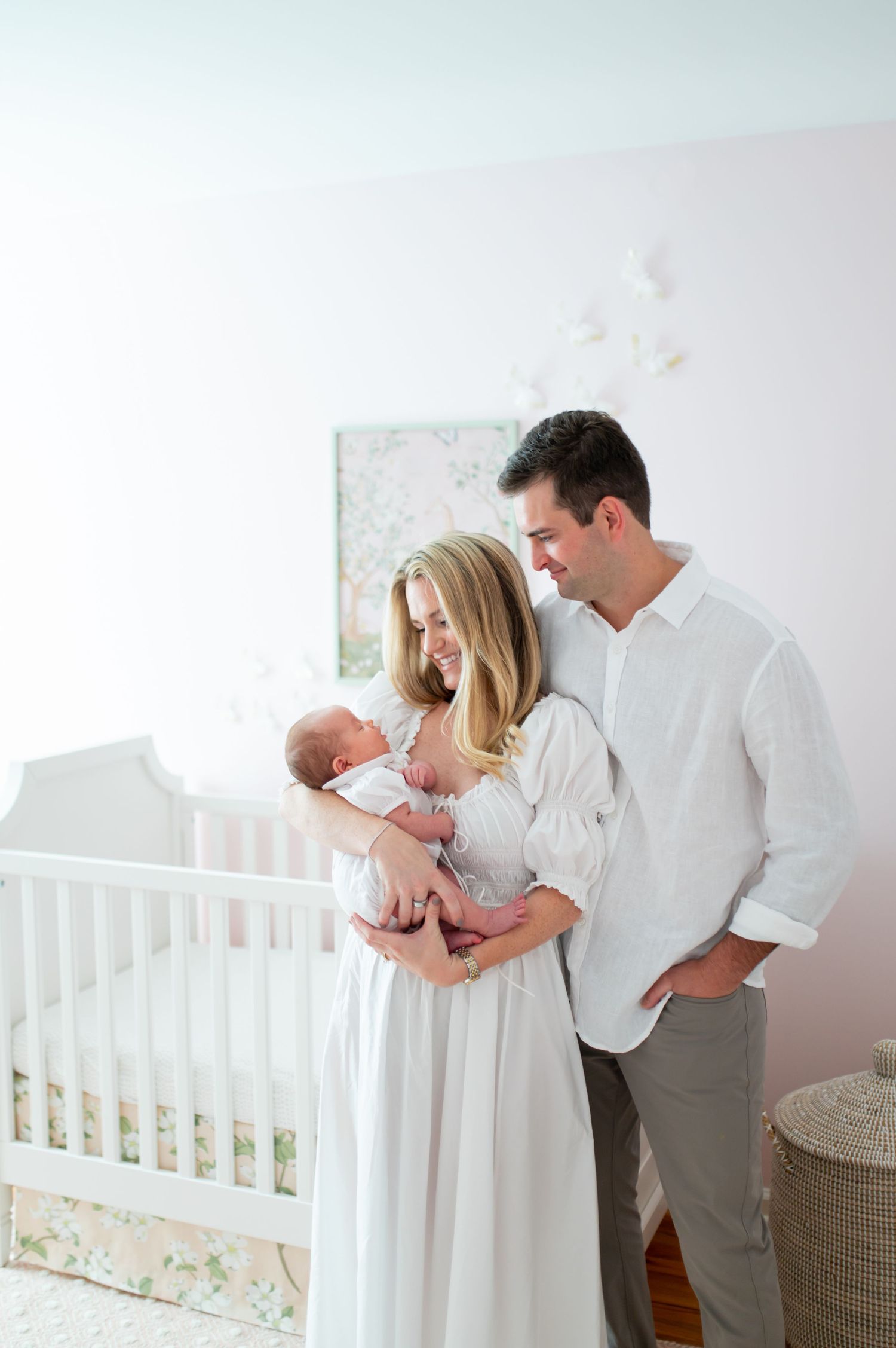 A bright white nursery room with a crib where family members gather in coordinated white and grey outfits.