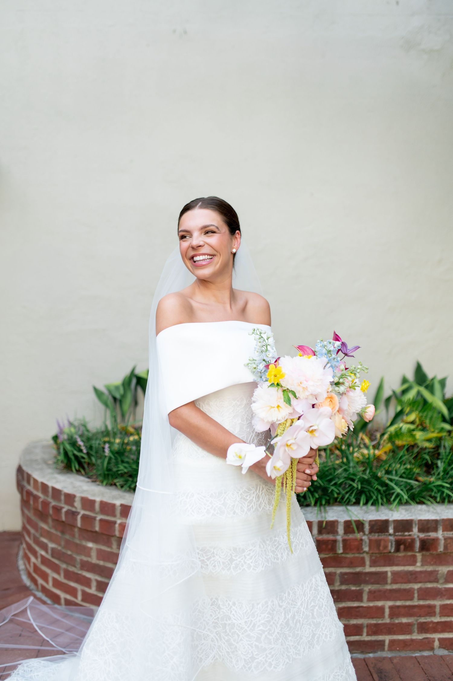 Radiant bride in off-shoulder lace wedding gown holding colorful spring bouquet outdoors.