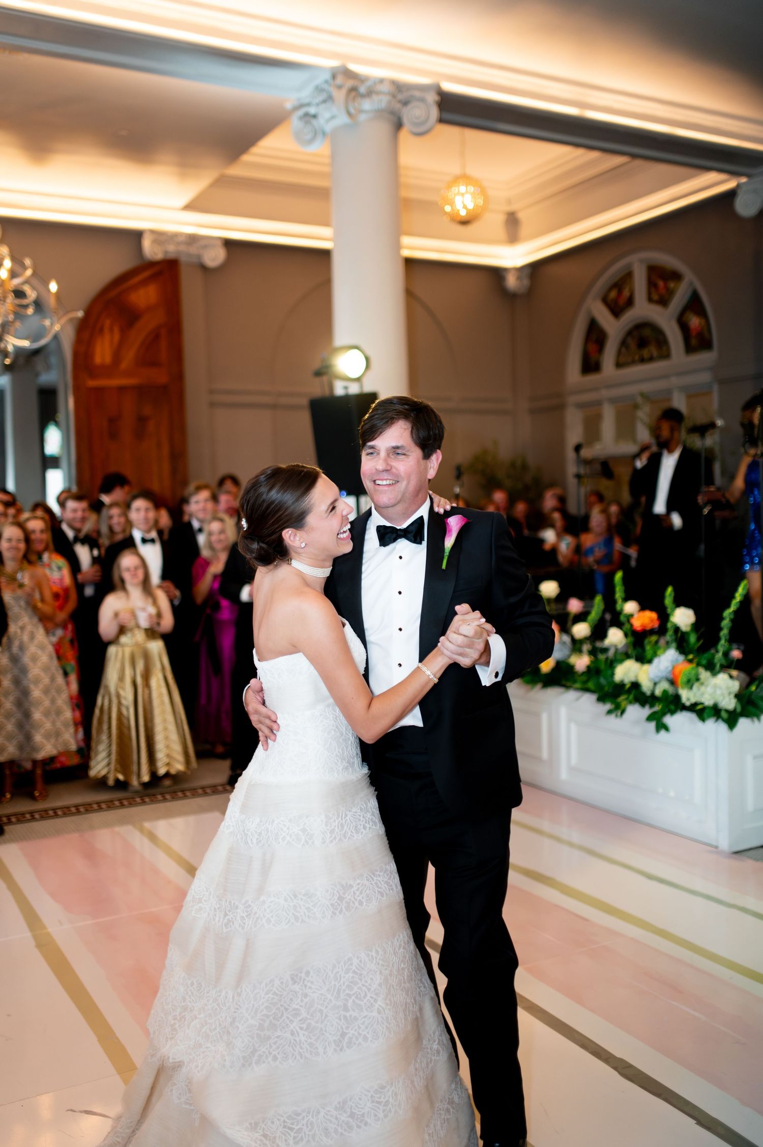 Smiling bride and groom share their first dance in an elegant ballroom surrounded by wedding guests.