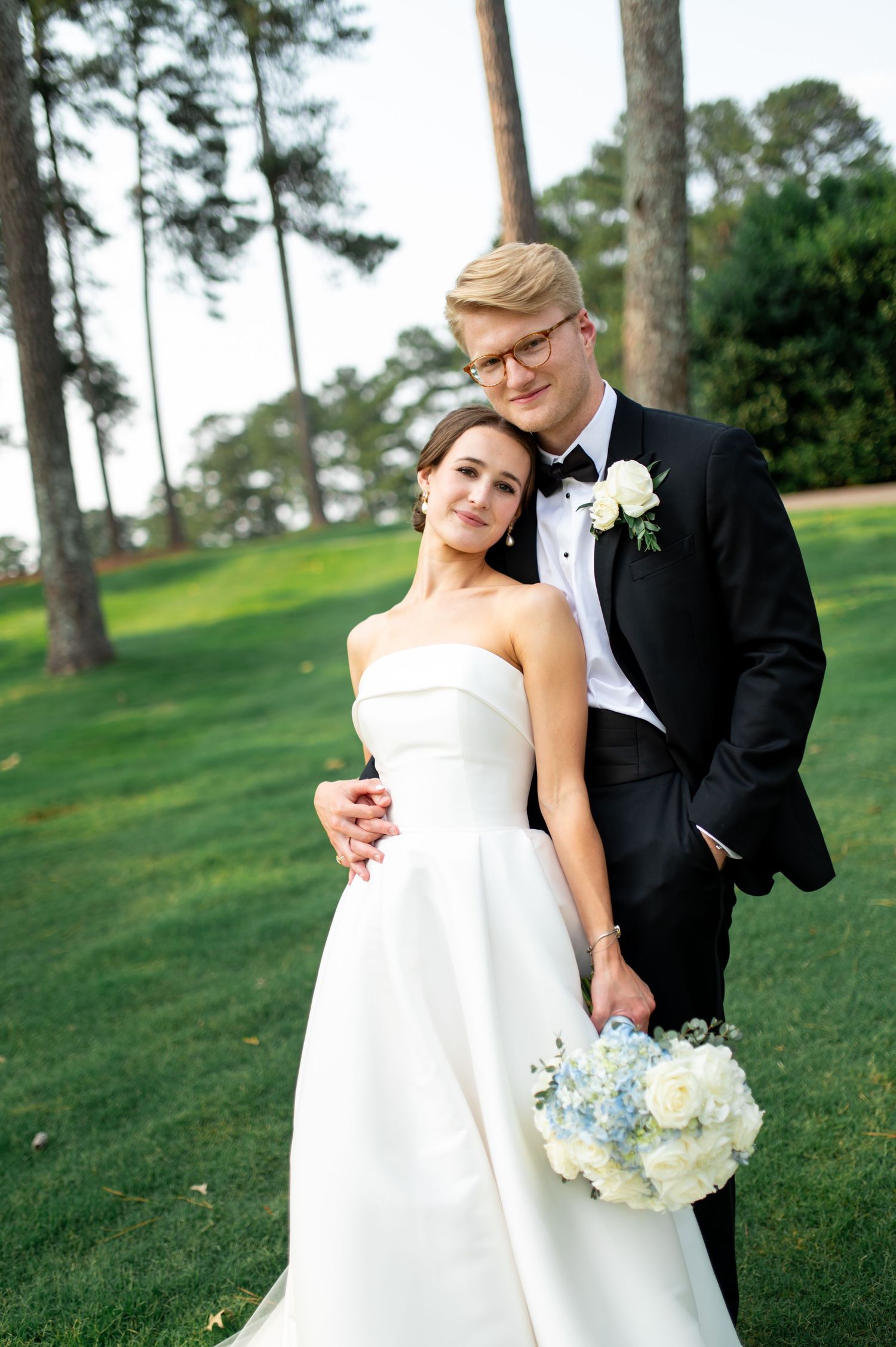 A romantic wedding couple poses together on a grassy hill with pine trees in the background.