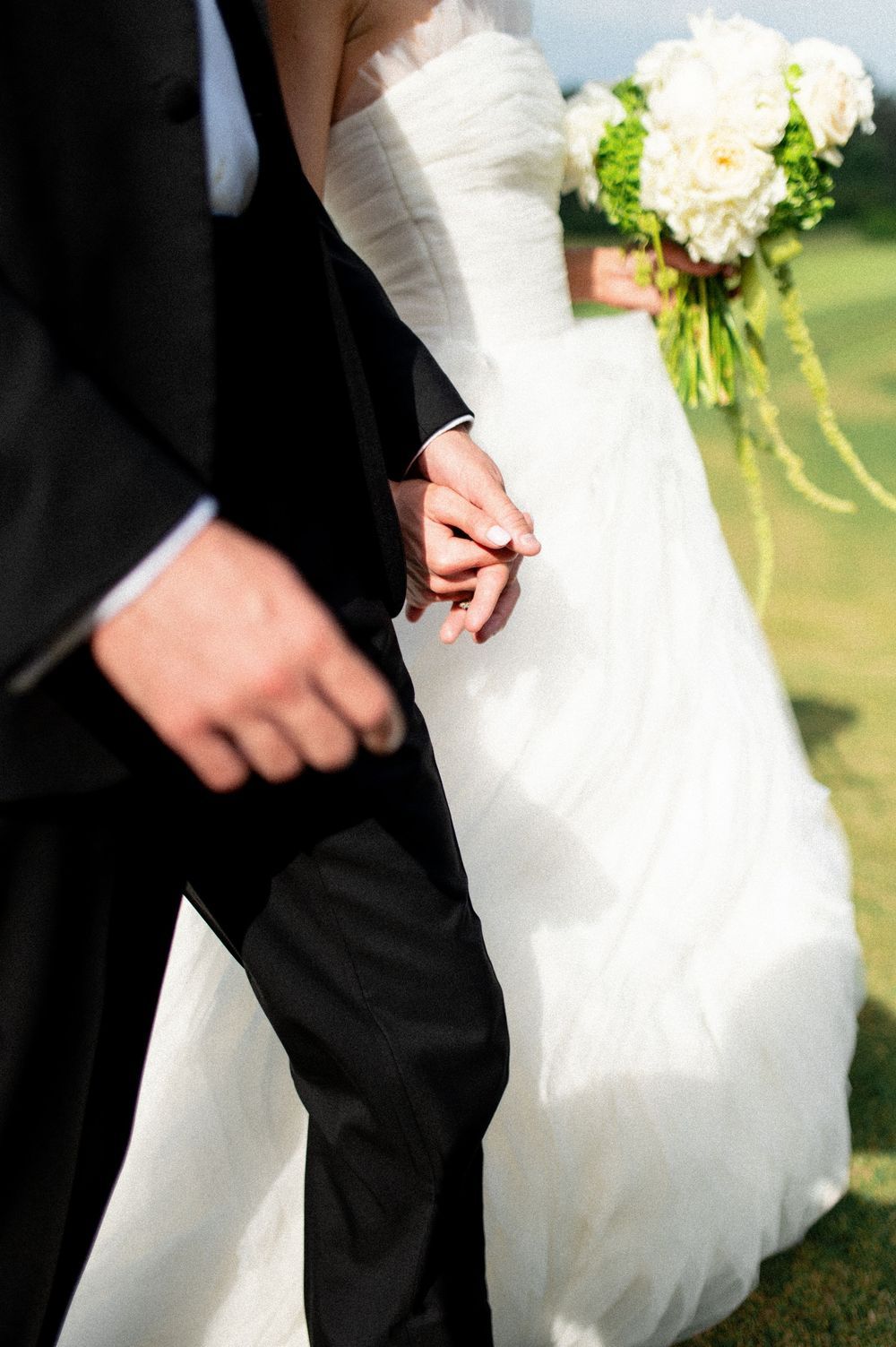 Bride and groom holding hands, bride in white gown holding green and white bouquet, groom in black suit.