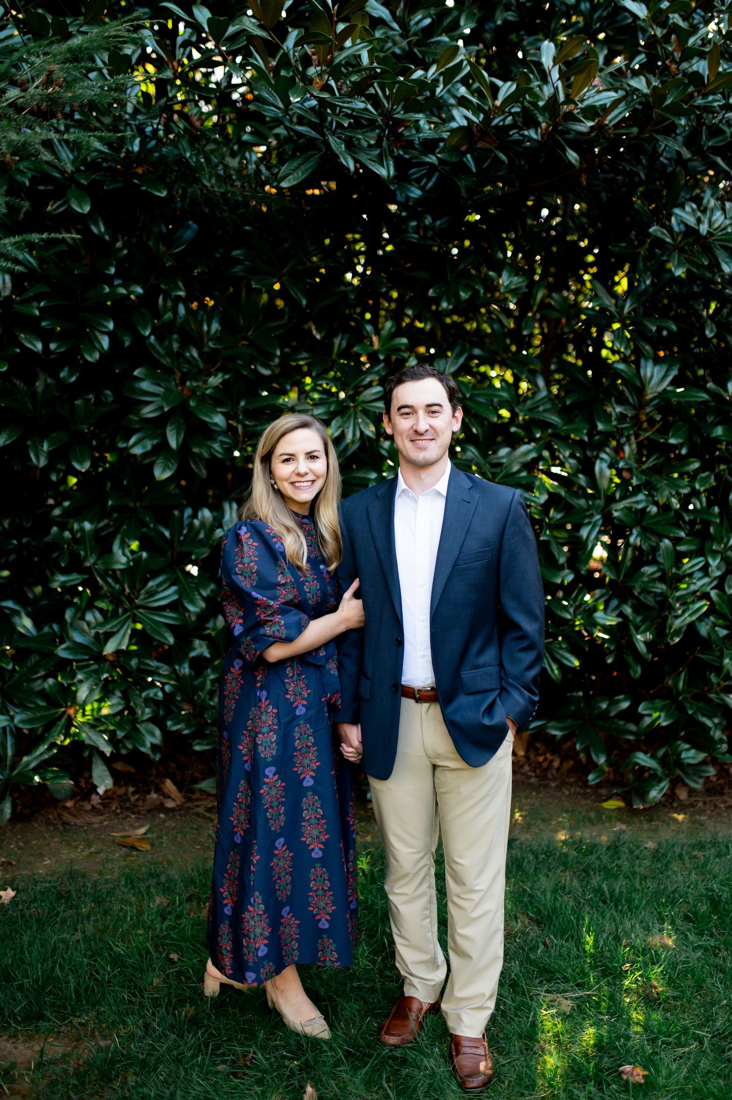 A smiling couple poses outdoors in front of lush green magnolia trees, dressed in smart casual attire.