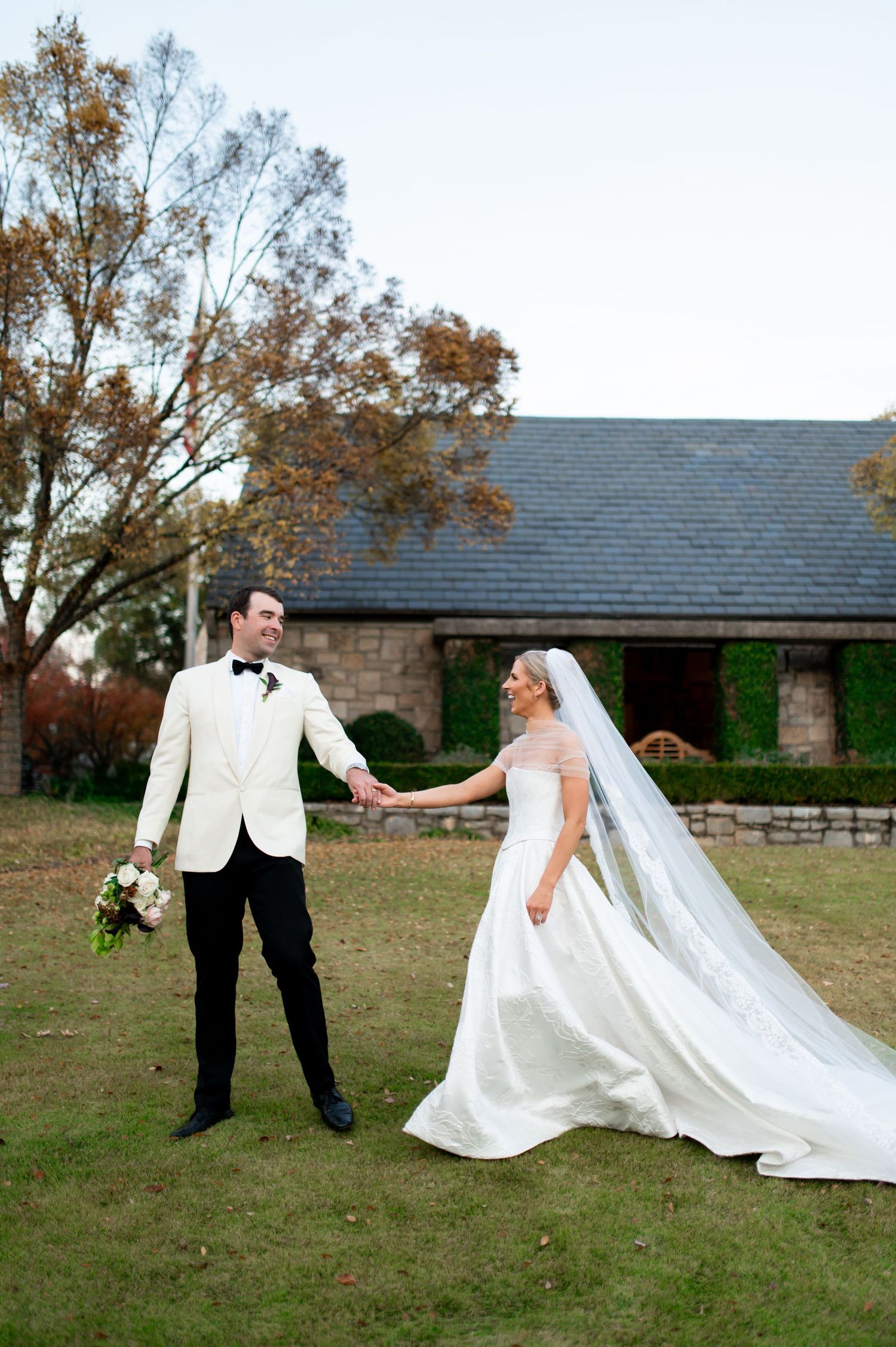 Wedding couple playfully posing with flowing dress on grass lawn near stone building.
