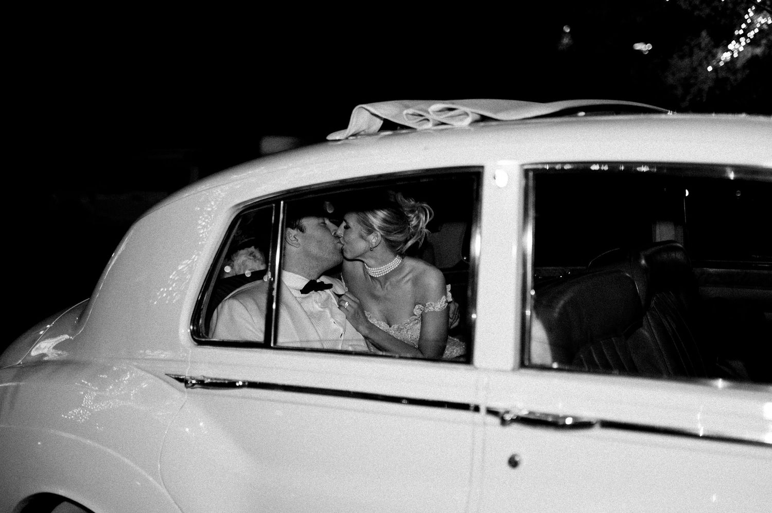 Classic white car departing with newlyweds after wedding celebration at night.