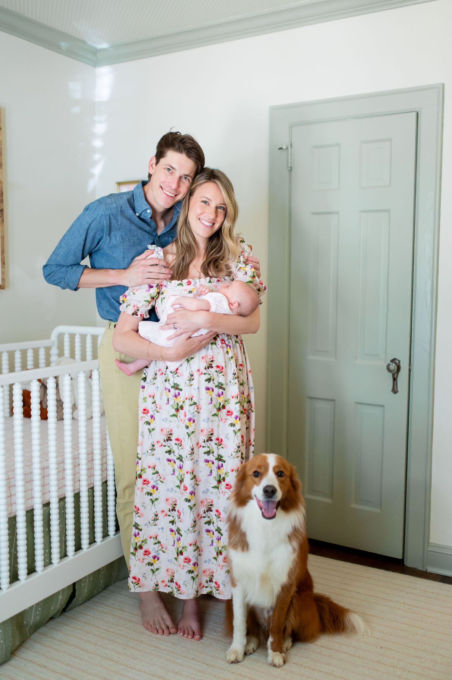 A couple stands in a nursery with their newborn baby and brown and white dog.