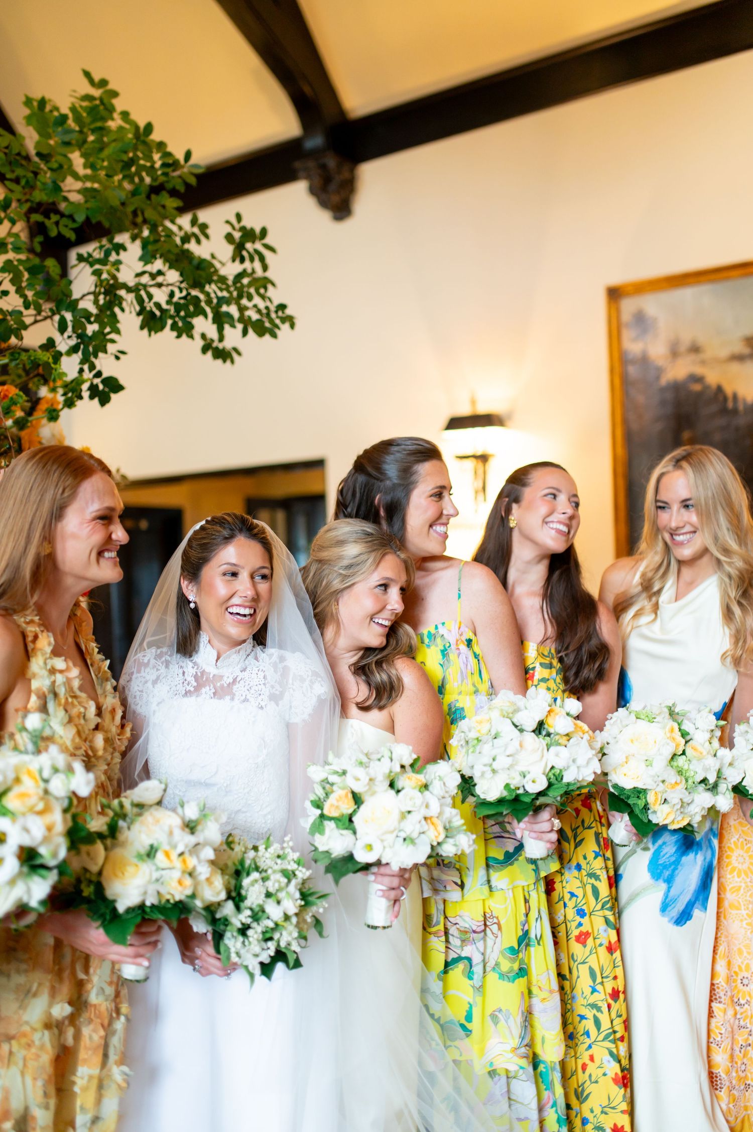 A wedding party in yellow floral and white dresses holds white bouquets while standing in a line indoors.