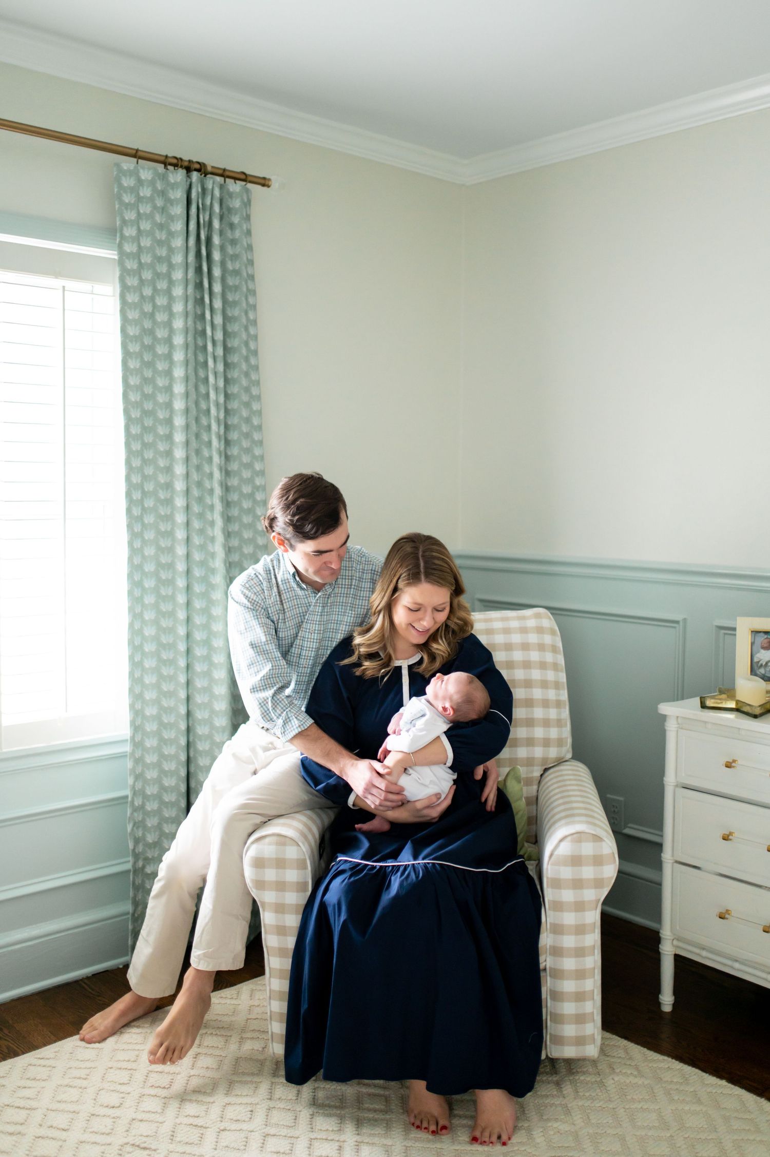 A cozy nursery scene with family members sitting in a striped armchair near blue curtains.
